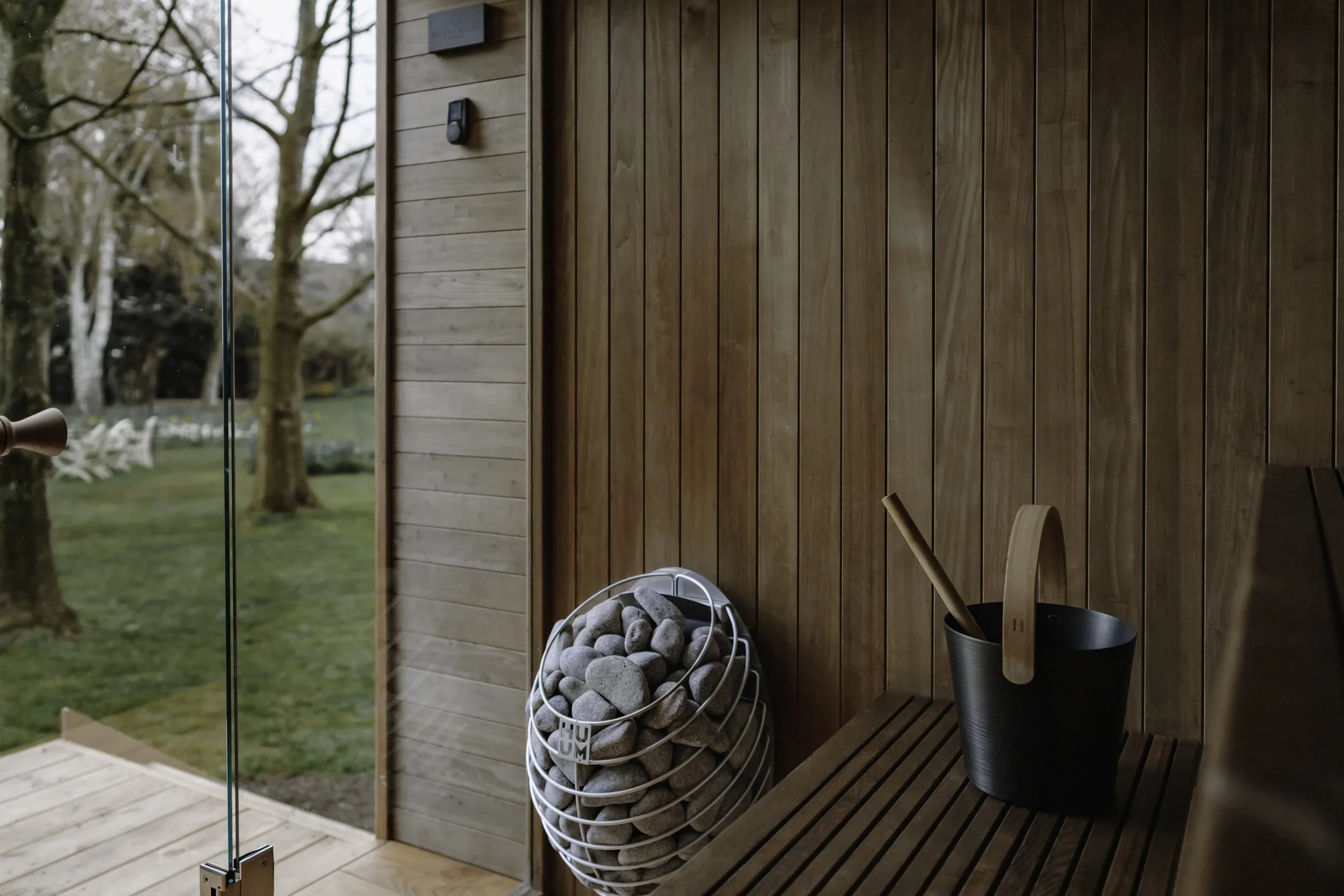 Interior of a sauna with wooden walls and a bench, a bucket with a ladle, and a wall-mounted electric HUUM sauna stove with grey stones. Outside the sauna, trees and green grass are visible through a glass door.