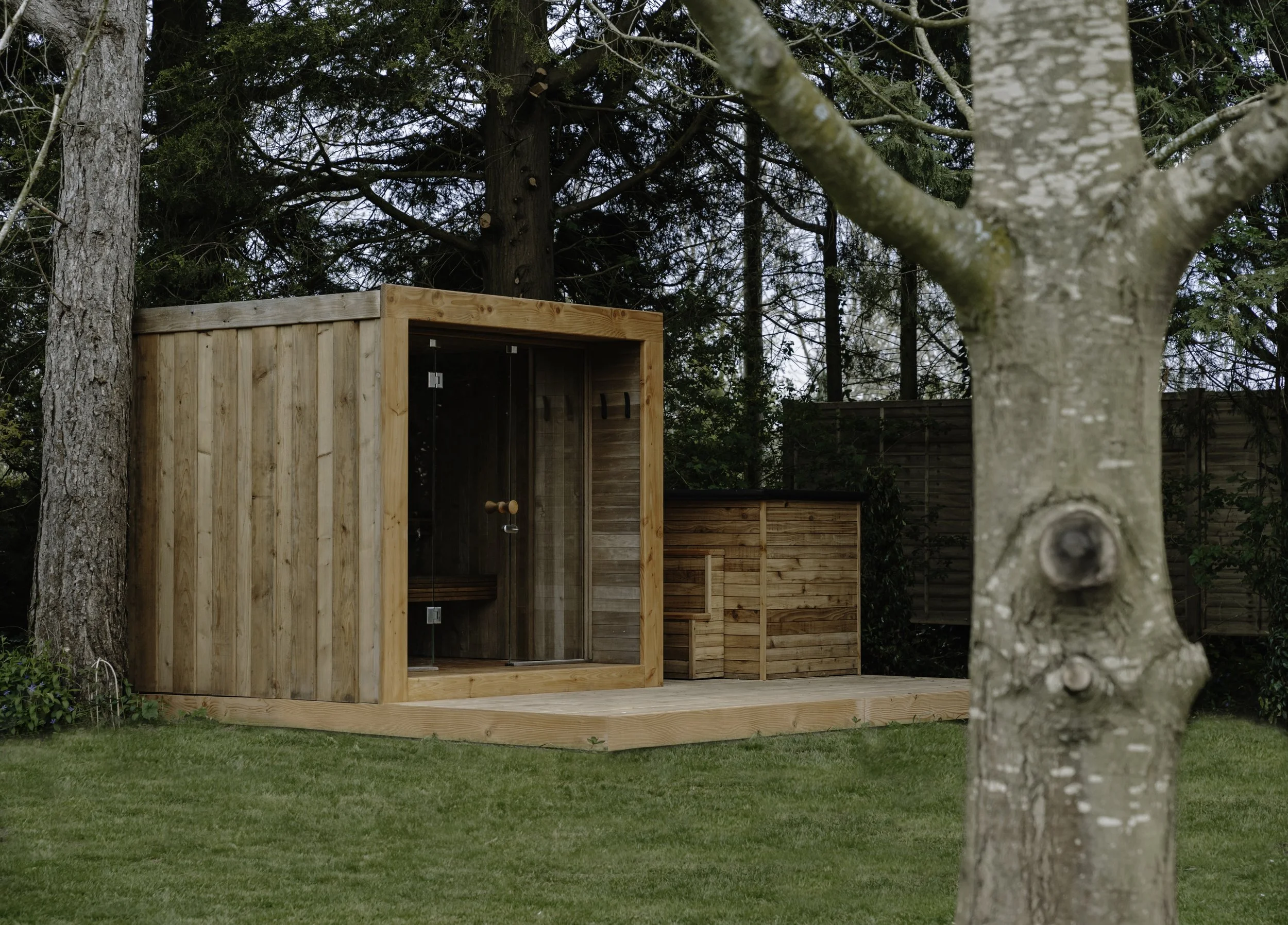 A backyard with trees and a lawn, a wooden plunge-pool and a wooden sauna on a deck with evergreen trees behind. 