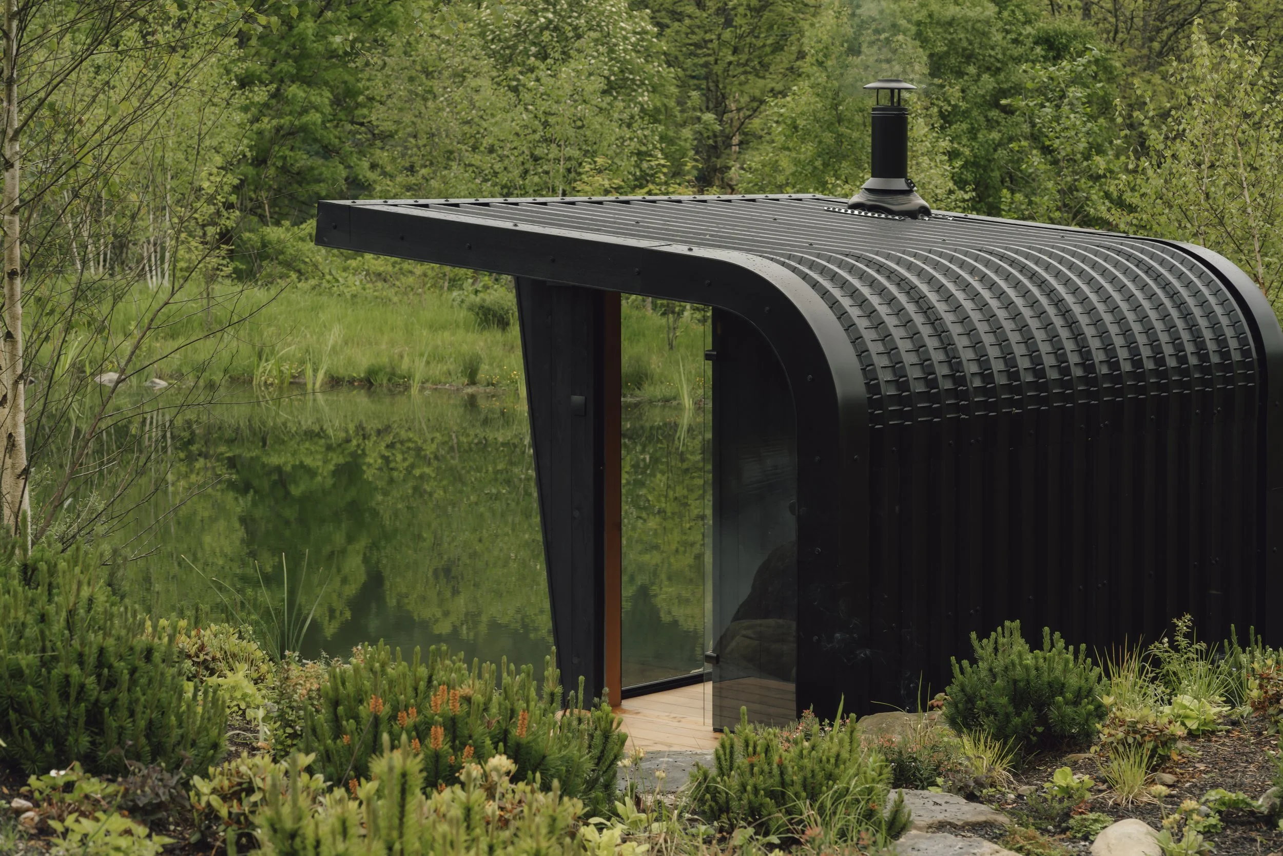 Small, modern black sauna with rounded roof and chimney, surrounded by green trees and plants, located near a calm lake or pond.