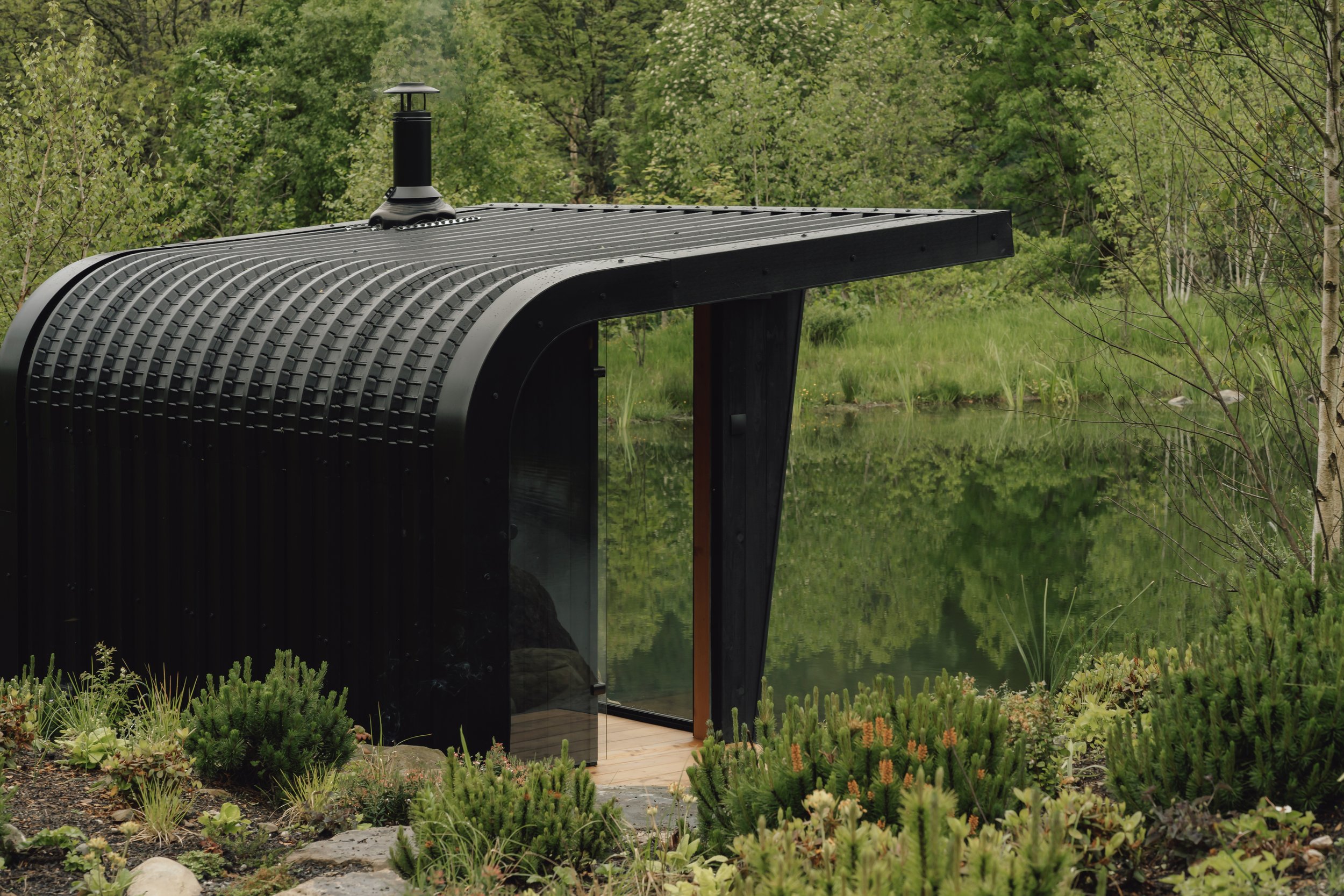 Modern black outdoor sauna with curved metal roof, glass door, and a chimney, situated in lush green garden near a natural swimming pond.