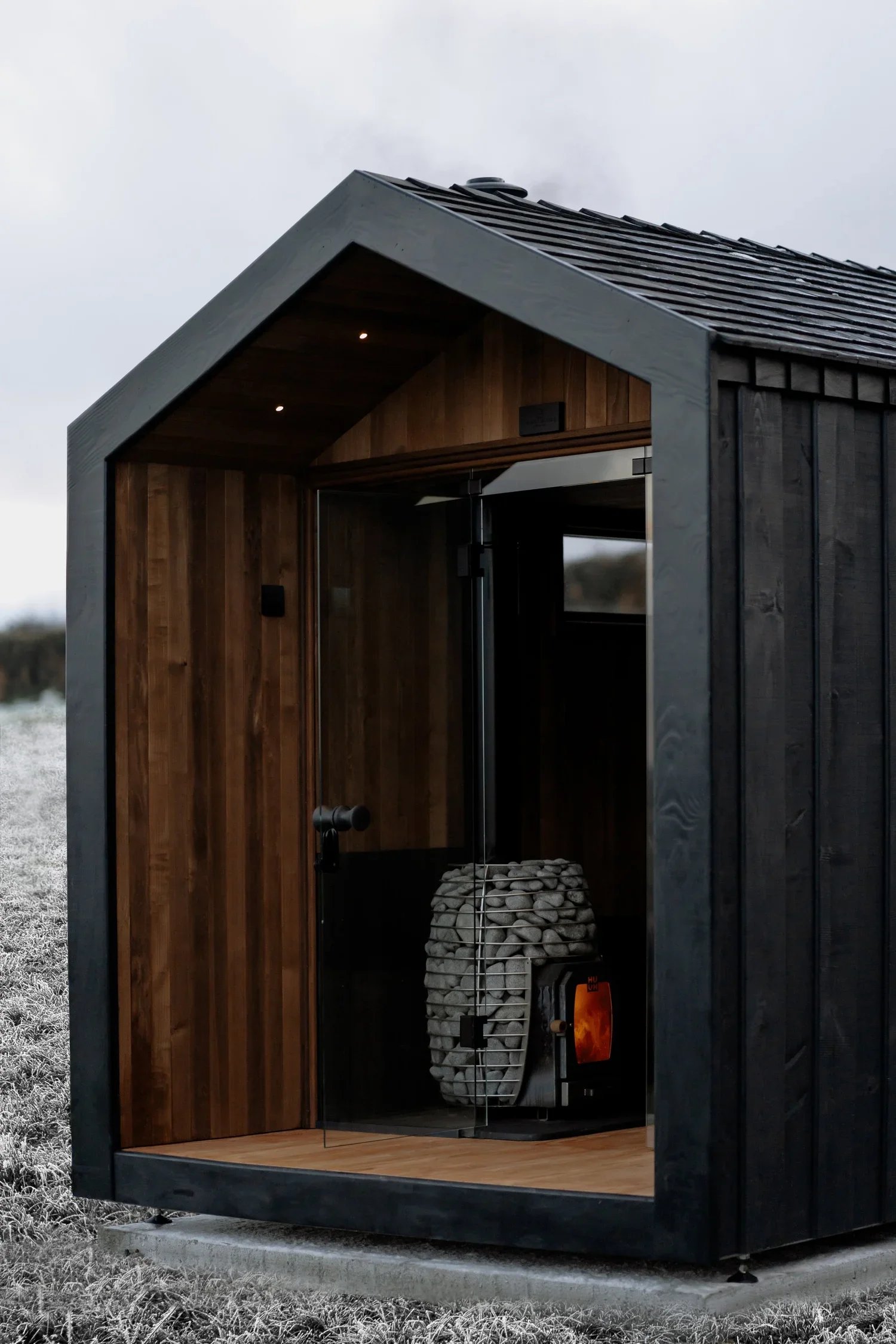 A black outdoor sauna with a glass door, wooden interior, and wood heater inside, situated outdoors on frosty grass.