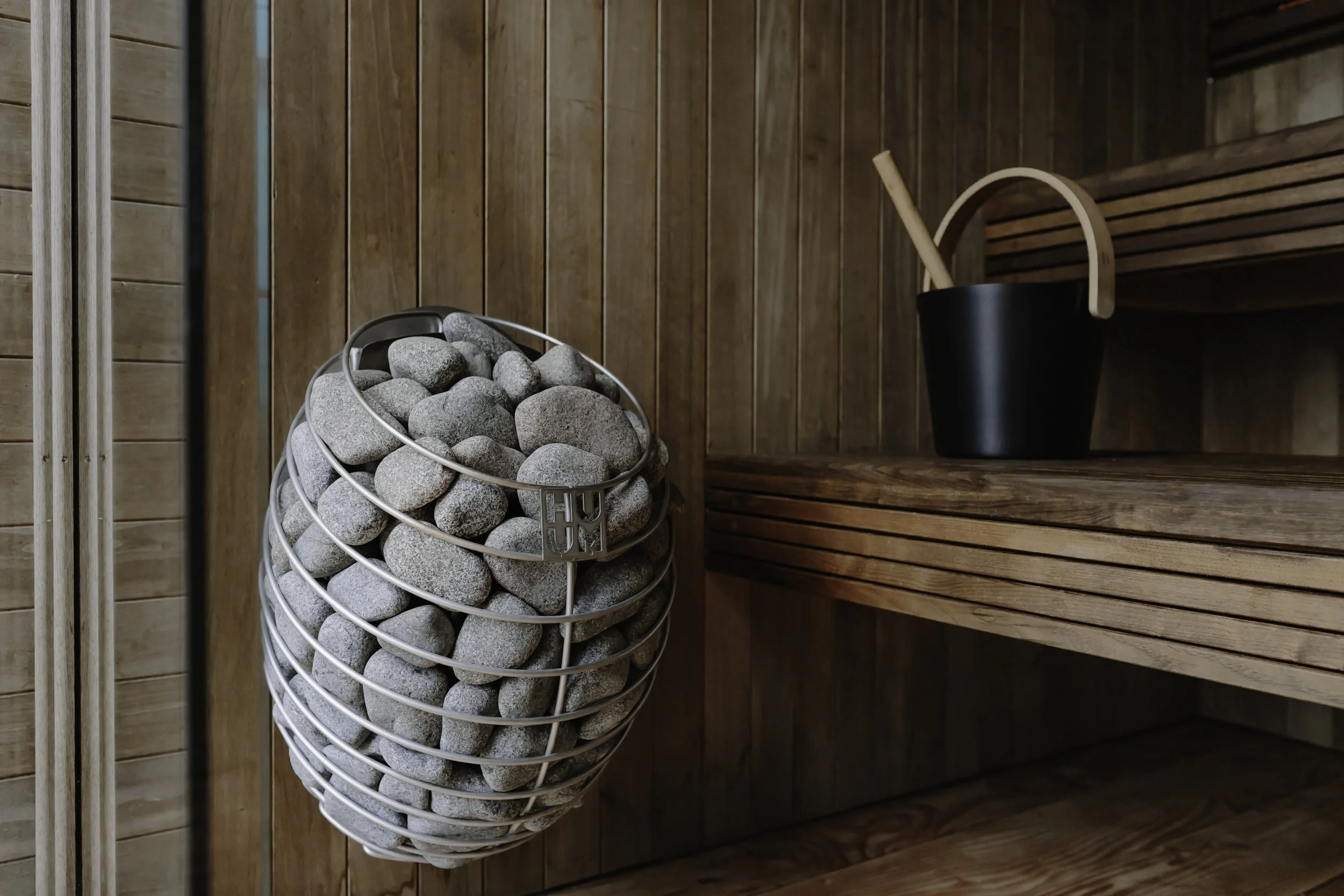 Interior of a warm wood-cladded sauna with an electric sauna of smooth gray stones and a black sauna bucket with a wooden handle.