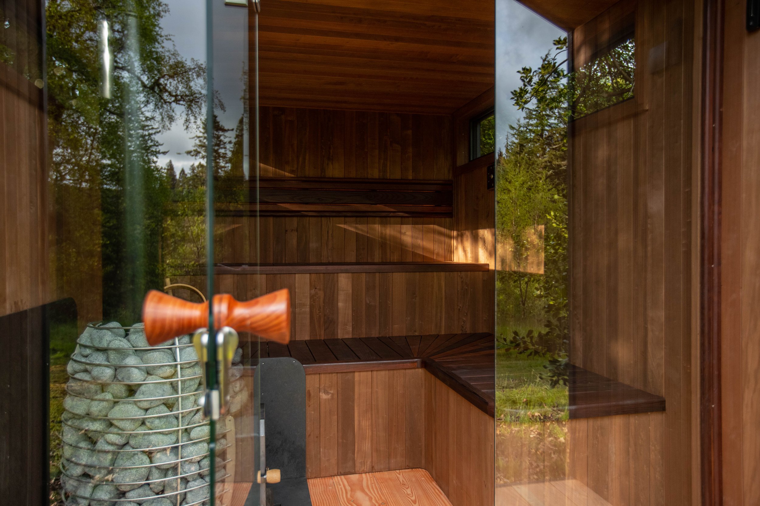 Inside a wooden outdoor sauna with glass door, wooden benches, and a heater with stones.
