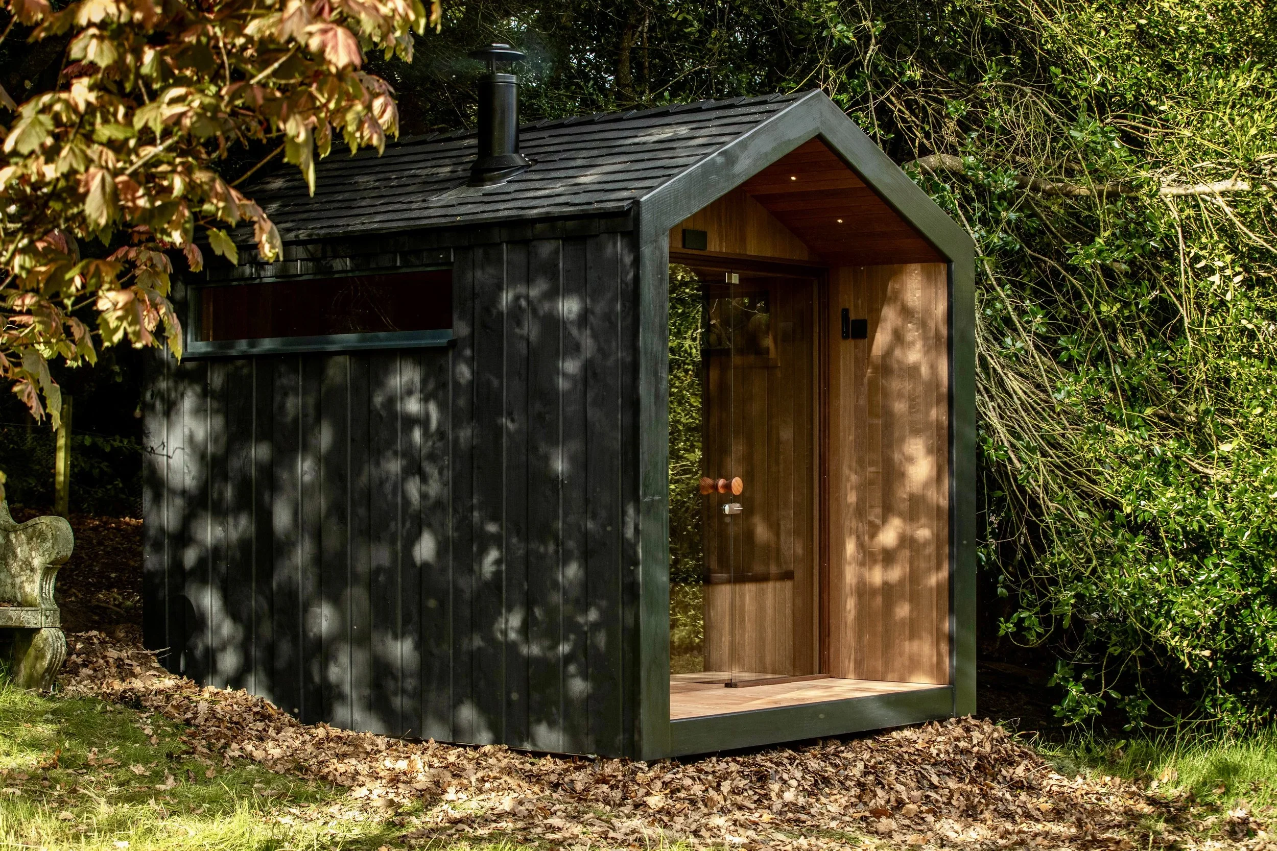Small black wooden sauna with a pitched roof, glass door, and a small landscape window, situated among trees and fallen Autumn leaves.