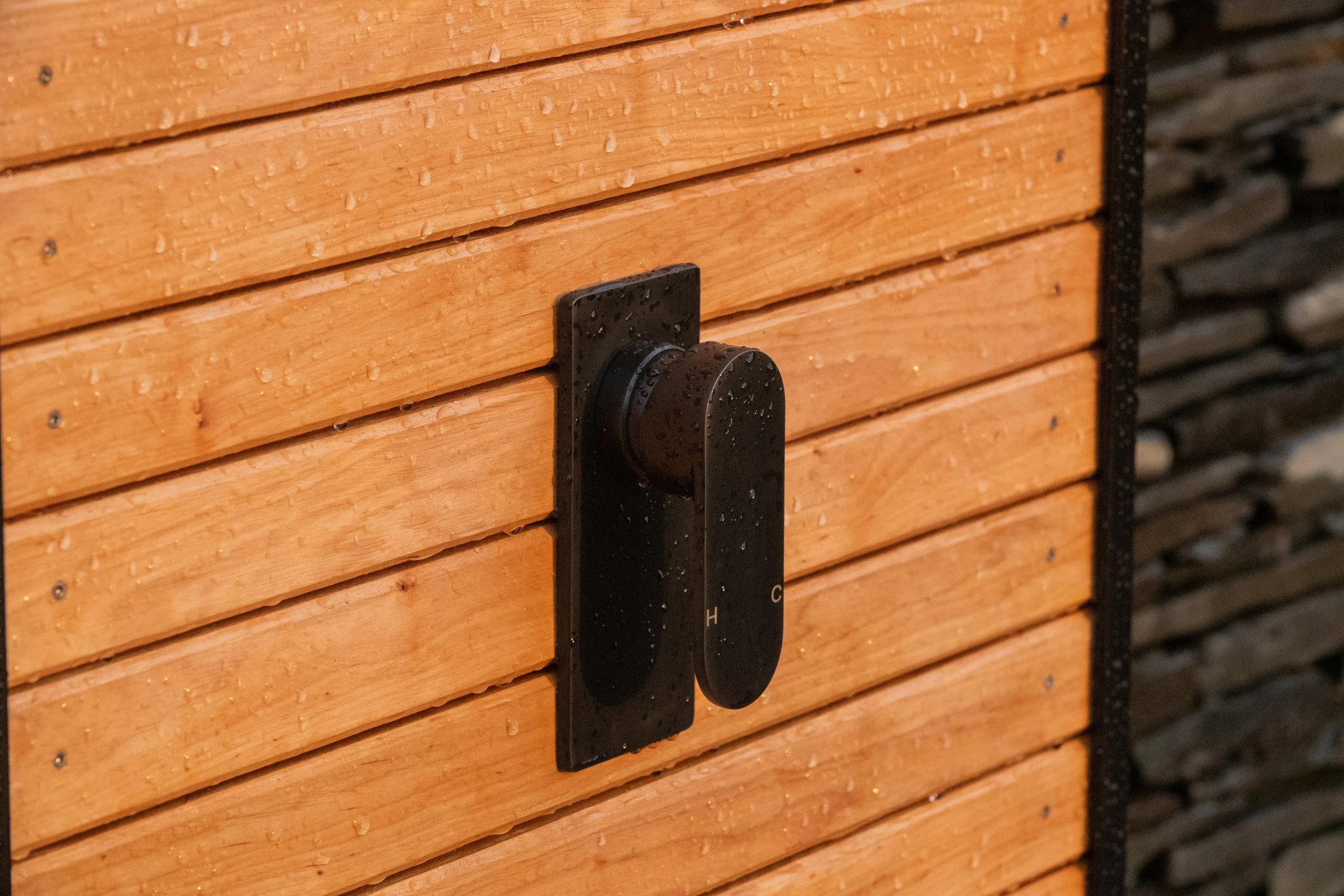A black thermostat mounted on a natural timber splash-back of an outdoor shower, with small water droplets.