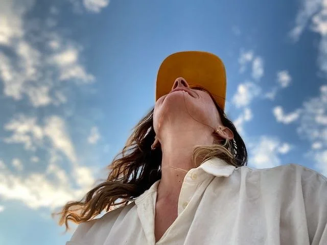 A woman wearing a white shirt and a yellow cap, looking up at the sky with clouds.