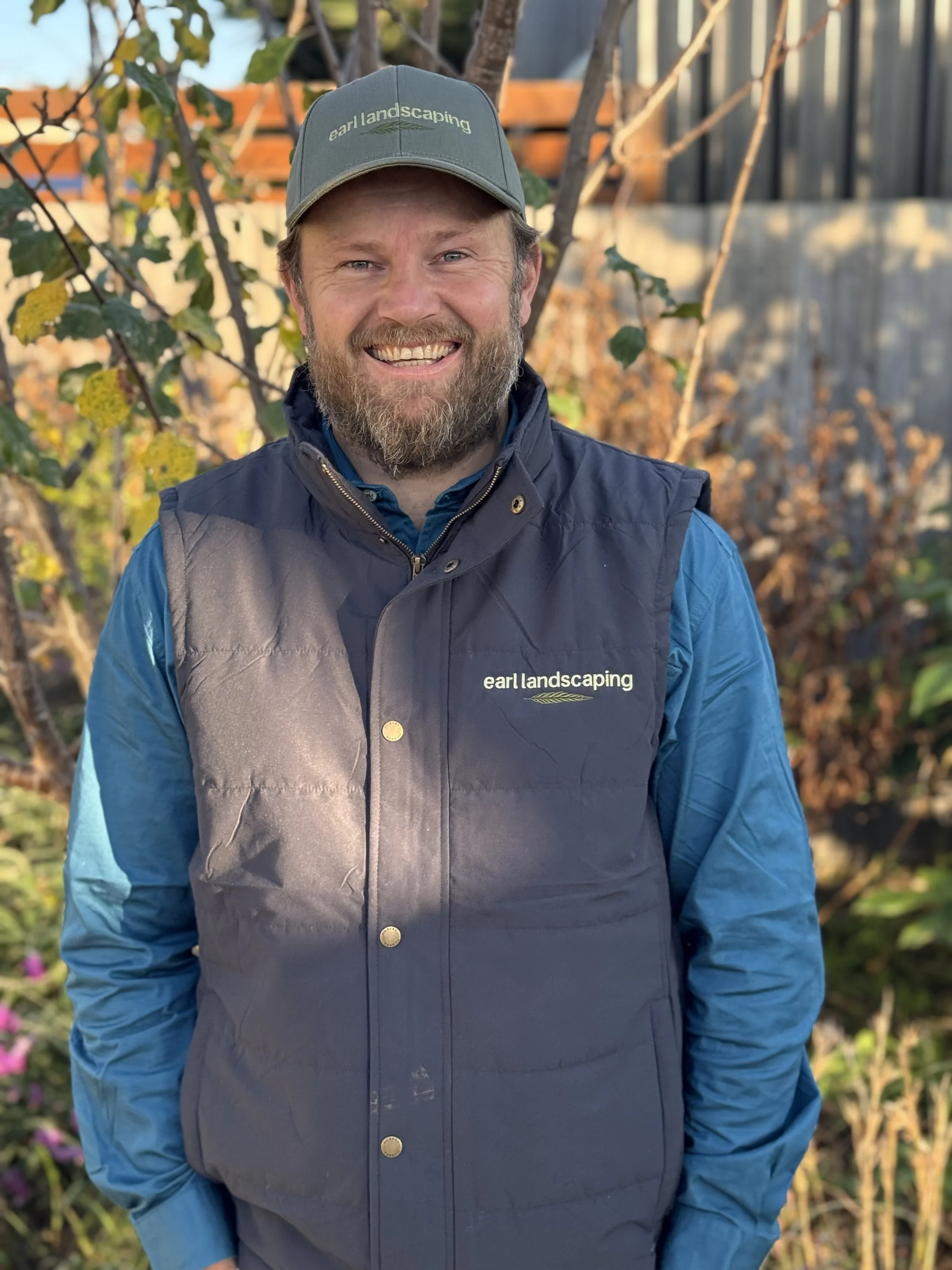 Portrait of Steve Earl smiling outdoors wearing a cap and a vest with 'earl landscaping' logo, standing in front of a garden with trees and some pink flowers visible in the background.