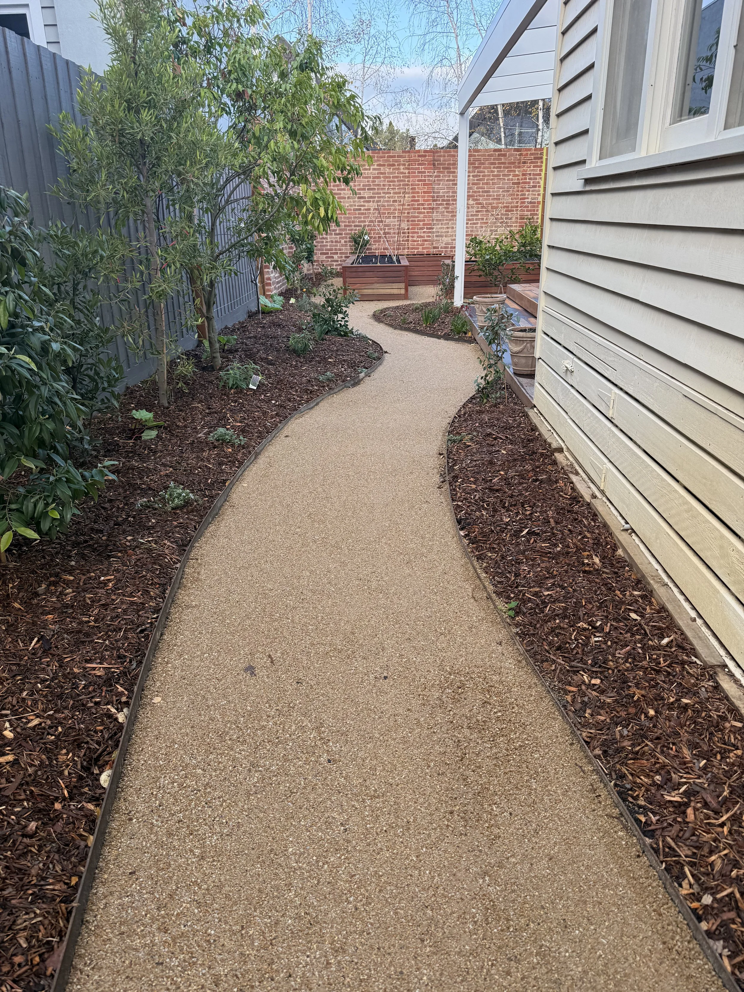 A curved garden pathway made of tan gravel runs through a backyard garden, flanked by plant beds with mulch, on the left side a large green shrub, and on the right side potted plants near a house with white siding, a brick wall in the background, and