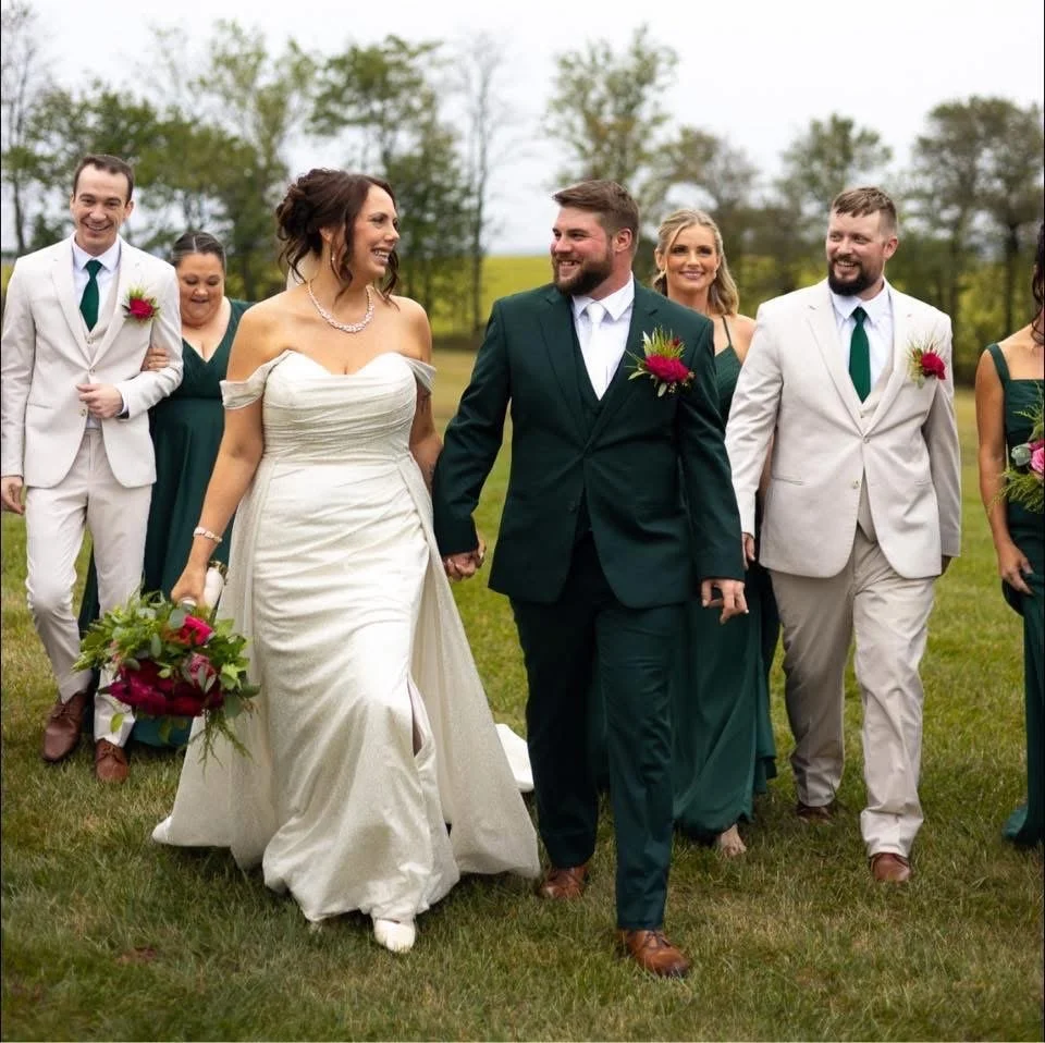 A wedding party outdoors with the bride and groom walking hand in hand, surrounded by bridesmaids and groomsmen in formal attire, on a grassy field with trees in the background.