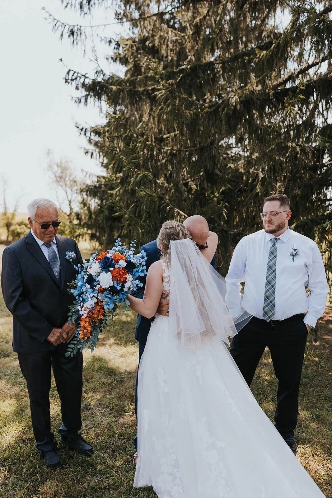 A wedding ceremony taking place outdoors under a large pine tree, with a bride in a white dress and veil, a groom in a dark suit, an officiant in a suit with sunglasses, and a man in a white shirt and plaid tie standing nearby. The bride is being kis