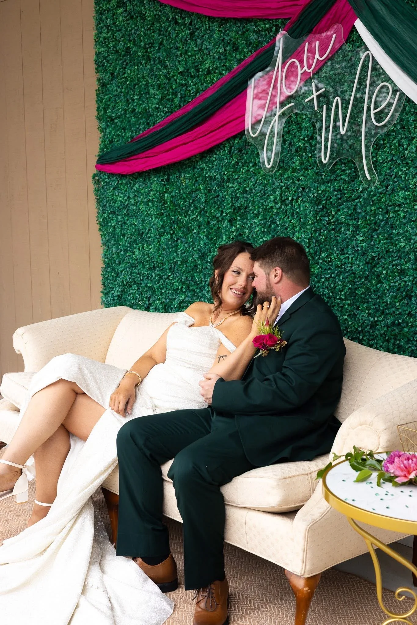 A bride and groom sitting on a beige couch, smiling and embracing at their wedding reception. Behind them is a green floral wall with pink and black drapery and a sign that says "You + Me" in white script.