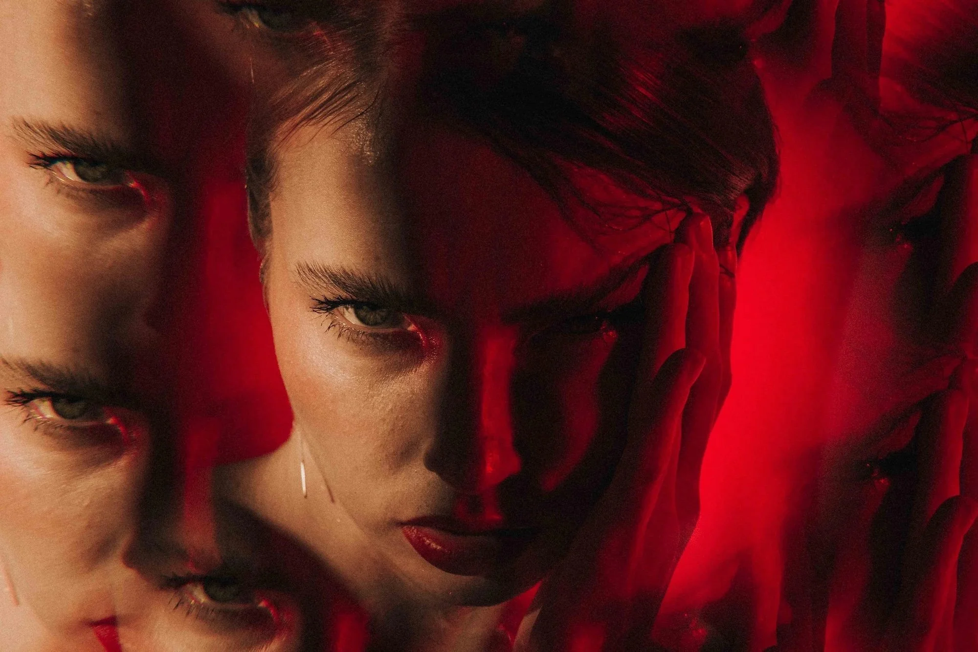 Close-up of a woman with pale skin and blonde hair, with a woman with dark hair and red clothing overlaid fused into her face, creating a layered effect.