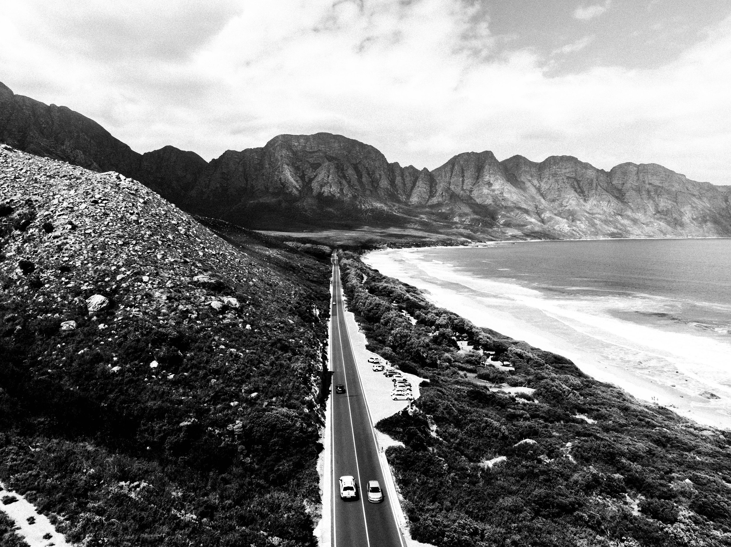 A black and white photo of a coastal mountainous landscape with a long road running through the middle, cars traveling along the road, and the ocean on the right side of the image.