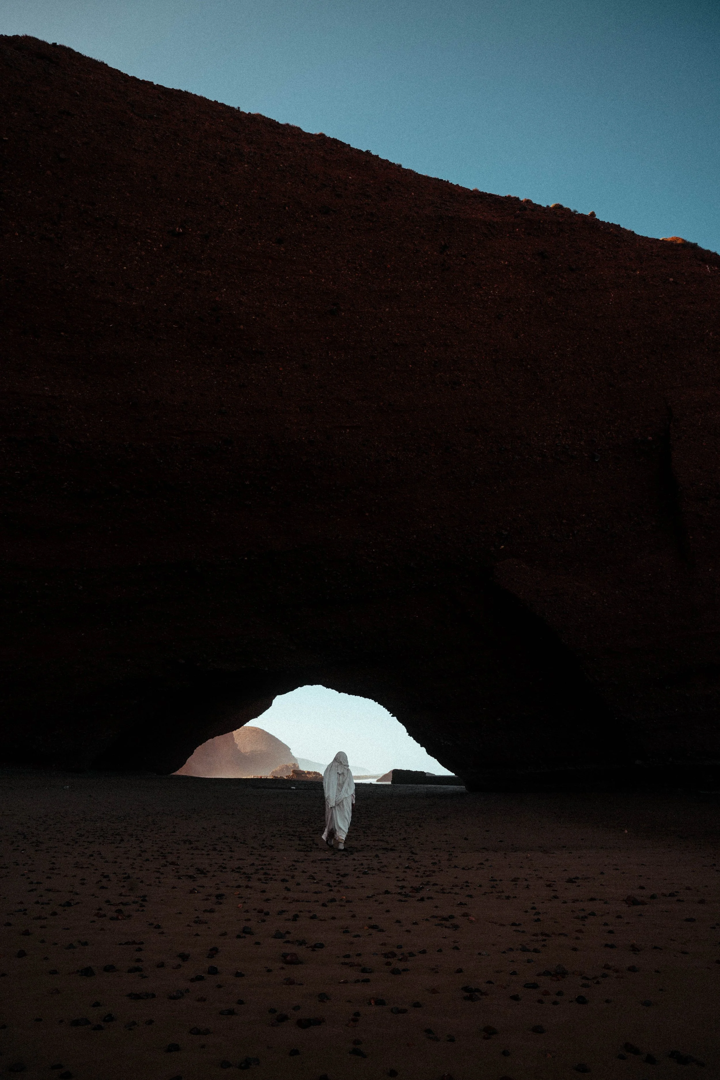 A person dressed in white walking through a dark, natural rock formation with a large arch and a view of the sky and distant mountains in the background during sunset or sunrise.