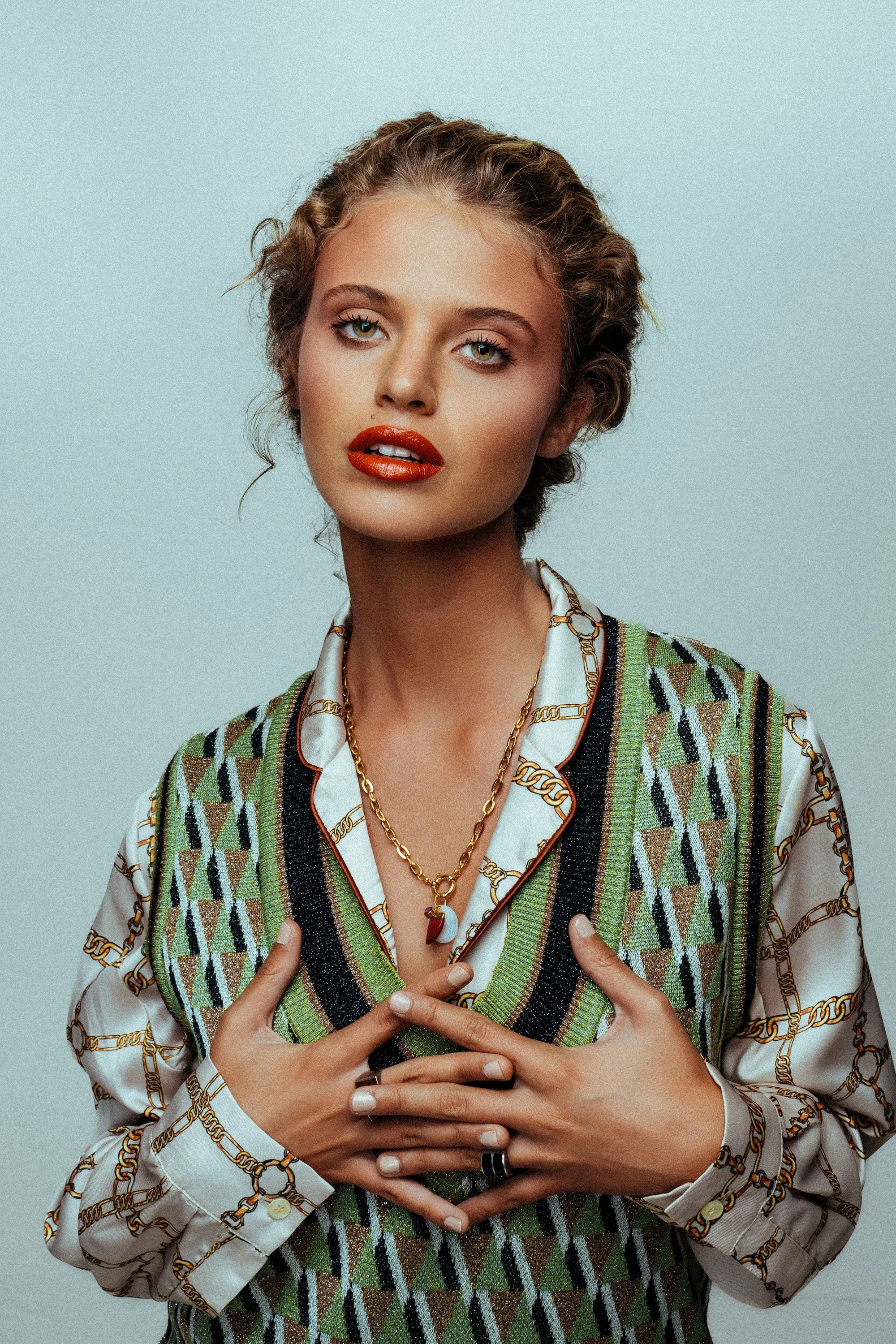 A woman with short wavy hair and red lipstick wearing a patterned blazer over a silk shirt, adorned with layered gold necklaces, holding her hands over her chest against a plain light background.