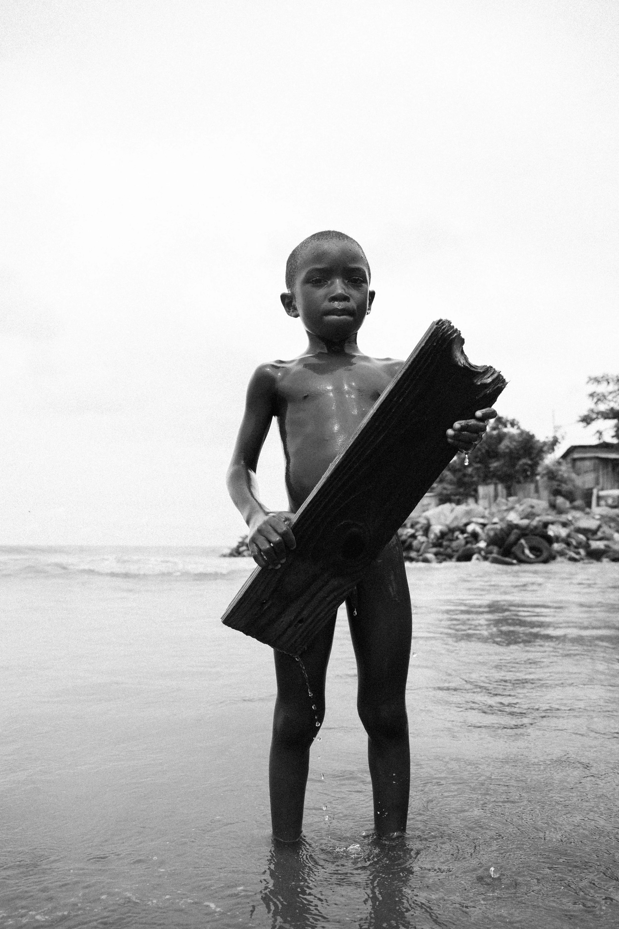 A young boy standing in the ocean holding a large piece of wood, with a serious expression, nearby rocks, and trees in the background.