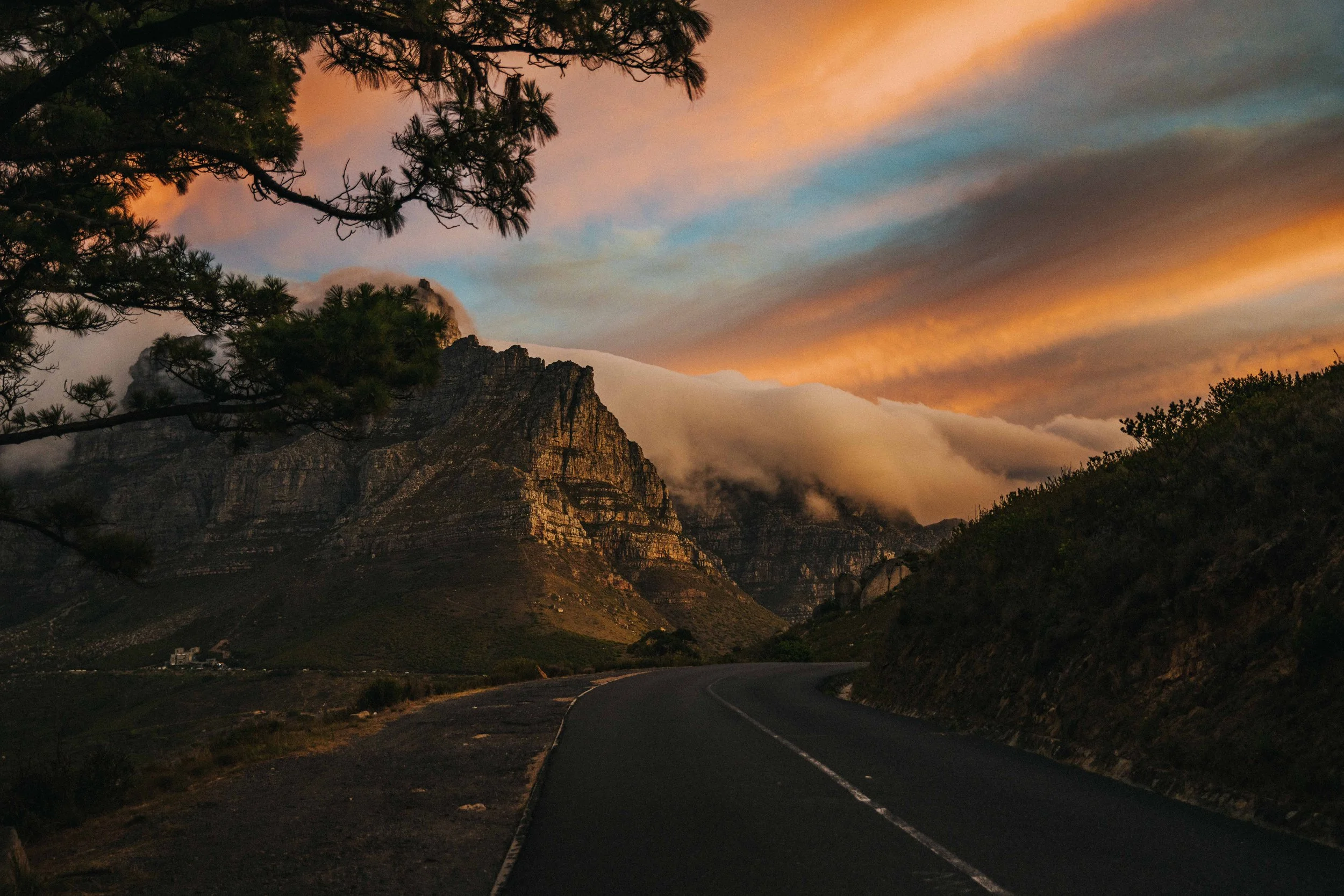A winding mountain road at sunset with tall rocky mountains in the background, and trees in the foreground, some with branches extending into the frame. The sky is painted with soft pastel hues of orange, pink, and blue.