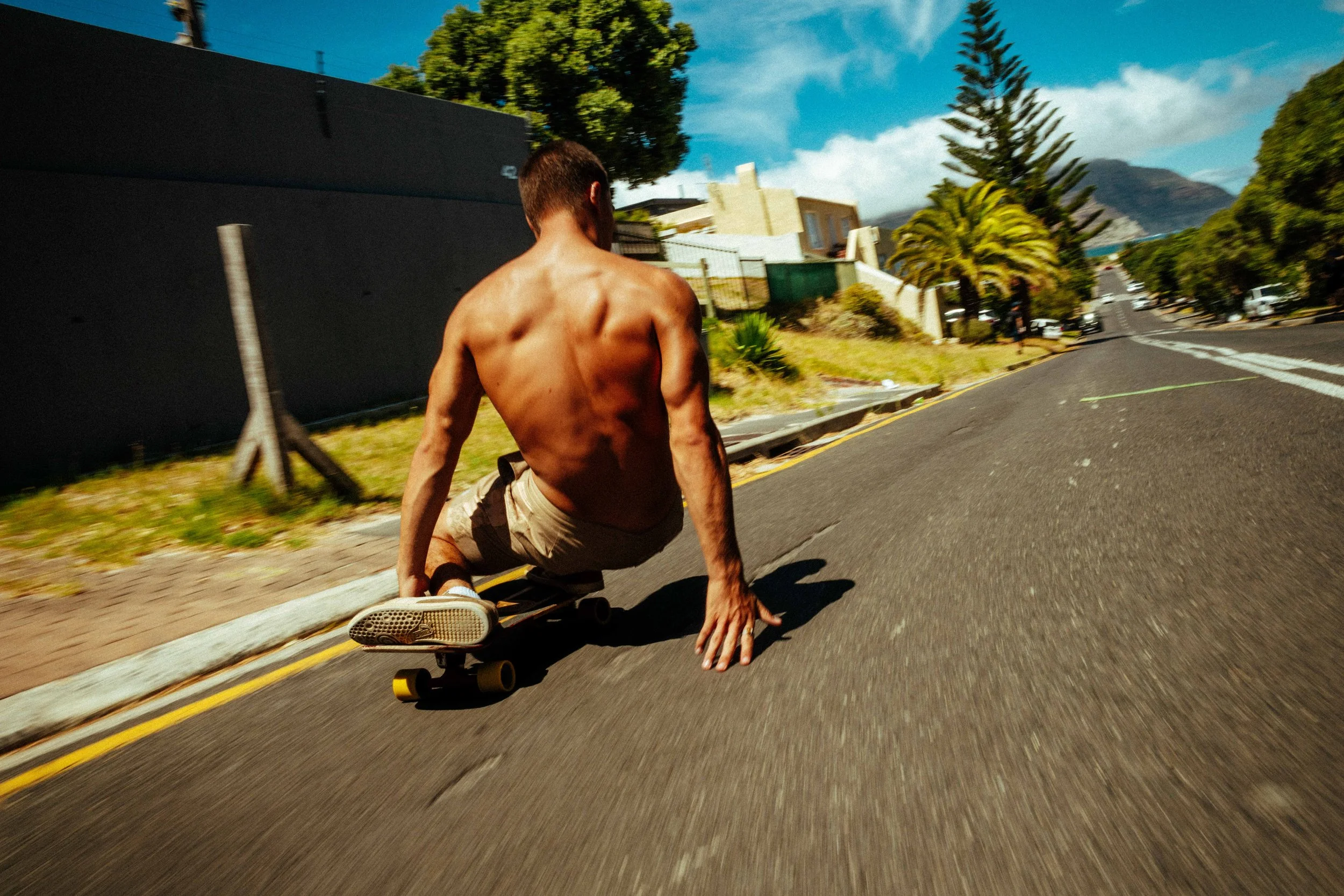 A shirtless man riding a skateboard on a city street, holding his left hand on the ground while balancing, with trees and buildings in the background on a sunny day.