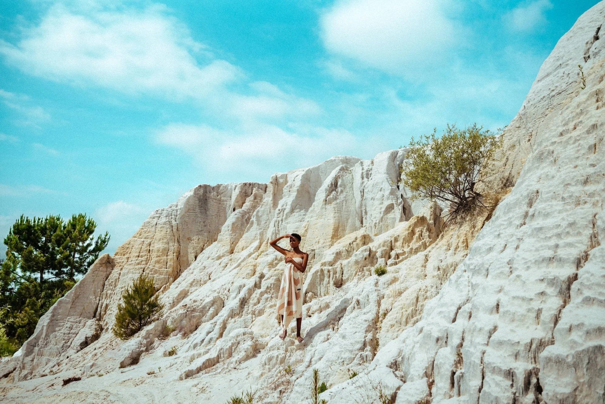 A woman standing on white rocky terrain with some green bushes and trees, under a partly cloudy blue sky.