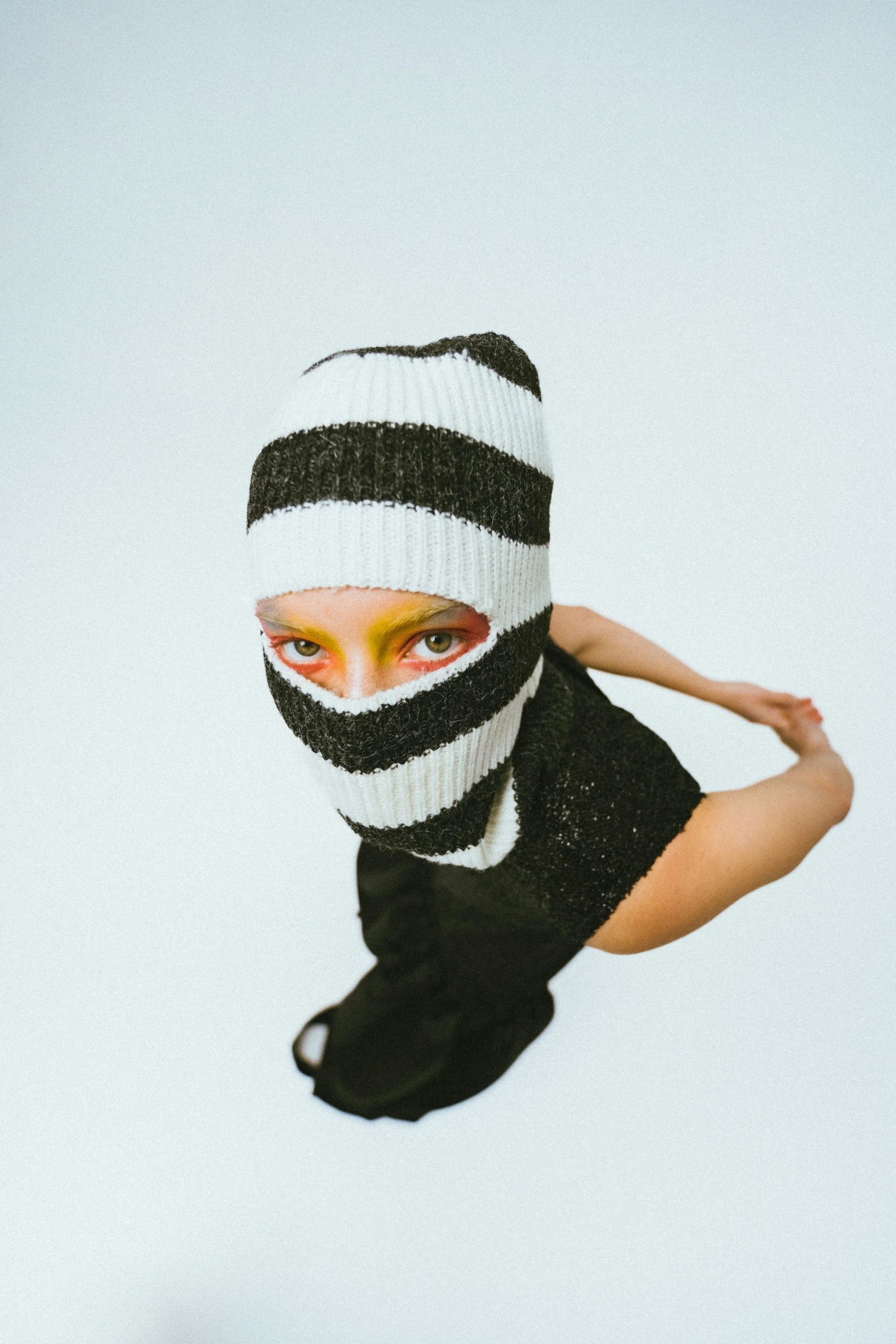 Person with face partially covered by a black-and-white striped knit mask, colorful eye makeup, wearing black clothing, posed with arms extended and leaning forward against a plain white background.