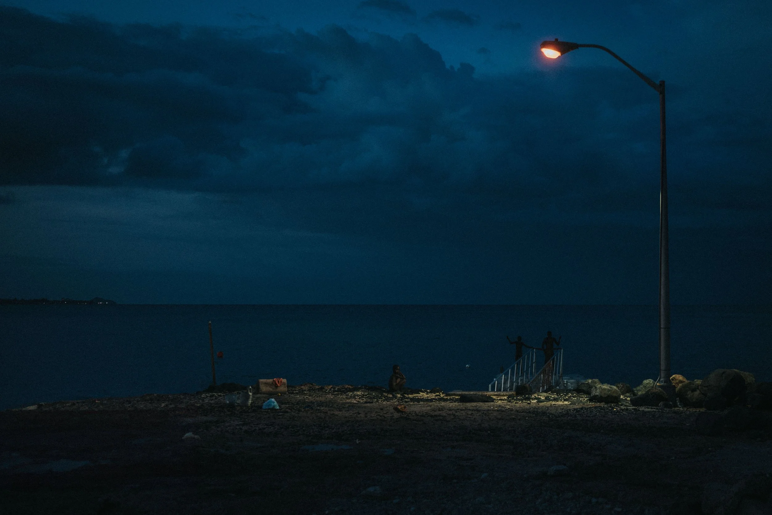 A dark evening scene by the water with a streetlamp illuminating the area. Two people are standing near a staircase leading down to the water, and the sky is filled with heavy clouds.