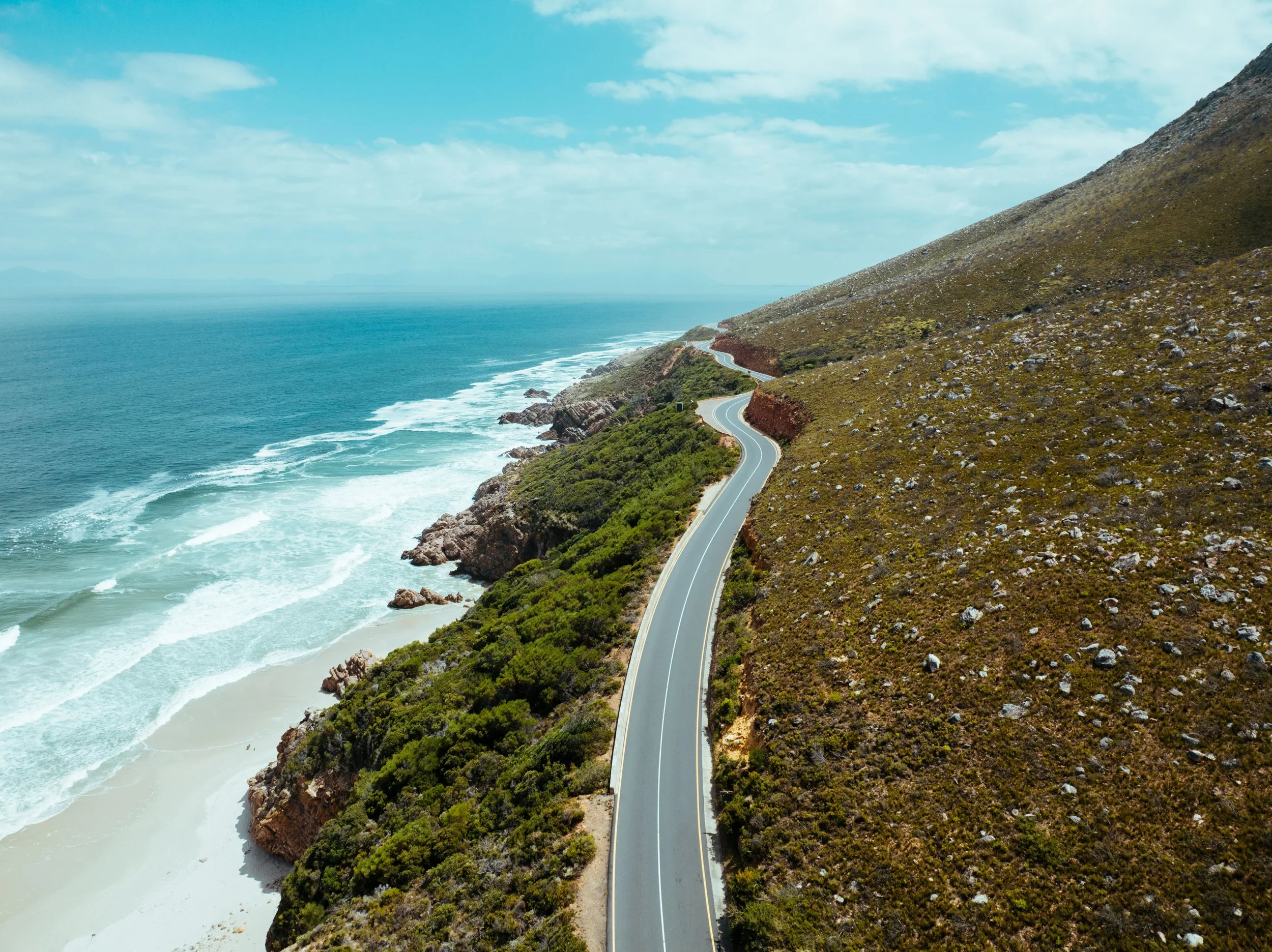 A winding coastal road runs along a hillside overlooking the ocean, with waves crashing on the beach below and a partly cloudy sky above.