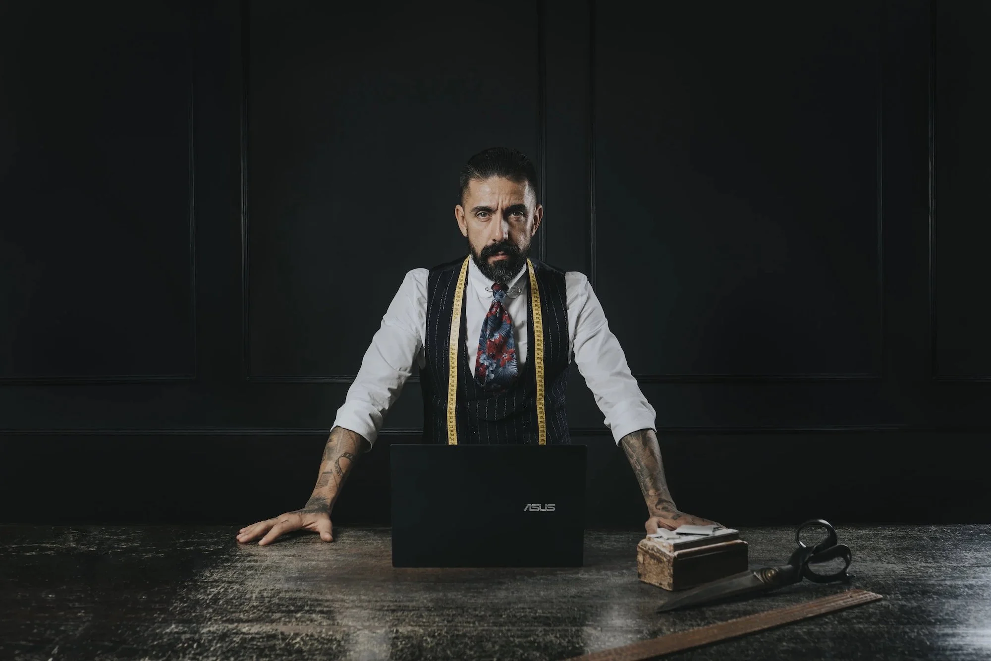 A man with a beard and tattoos, wearing a white shirt, black vest, and a colorful patterned tie, is sitting at a dark wooden table in a modern, black-walled room. He has a measuring tape draped around his neck and is leaning forward with his hands on