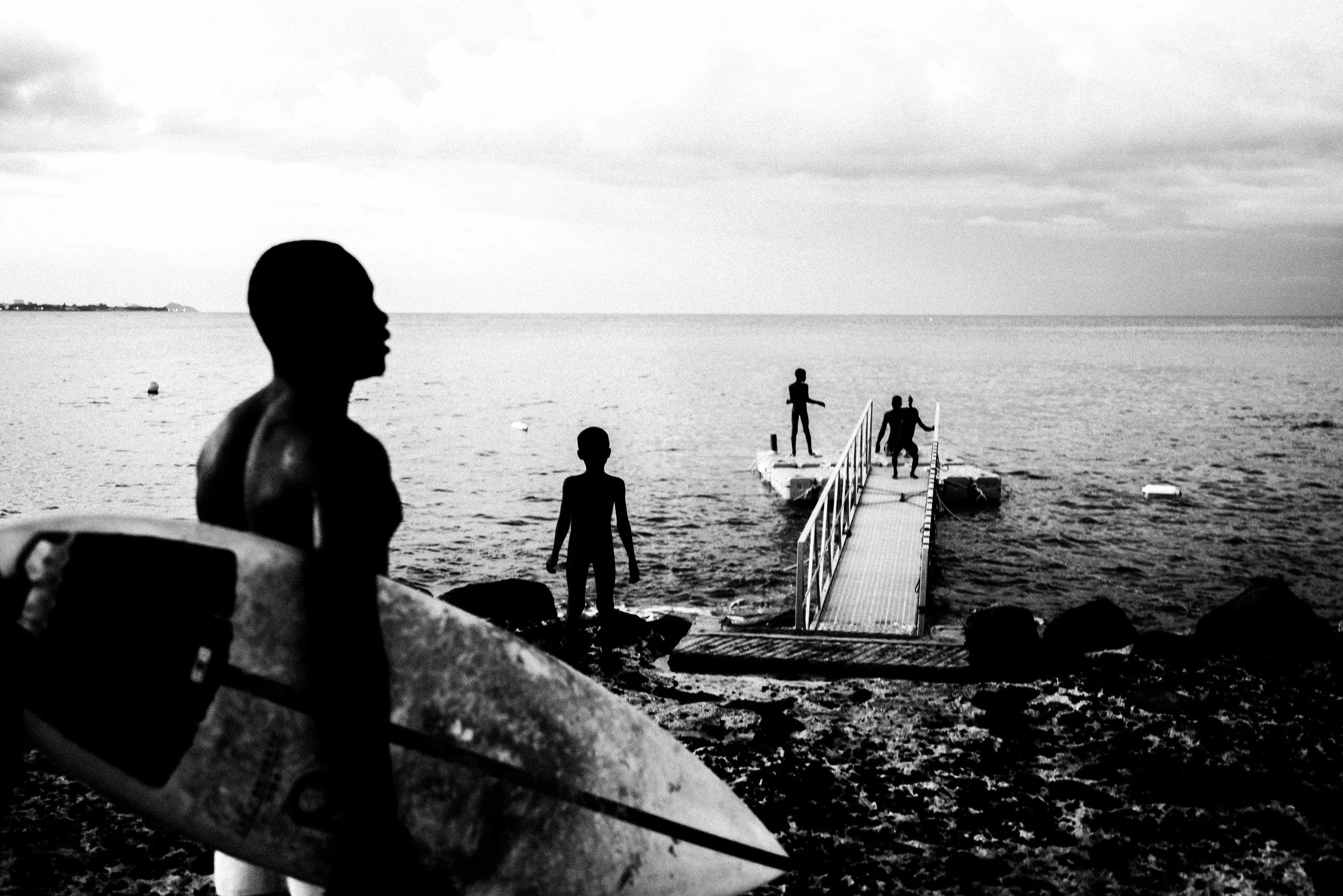 Silhouettes of people near a dock by the water, with one person standing on the dock and others in the water, under cloudy skies in a black and white photo.