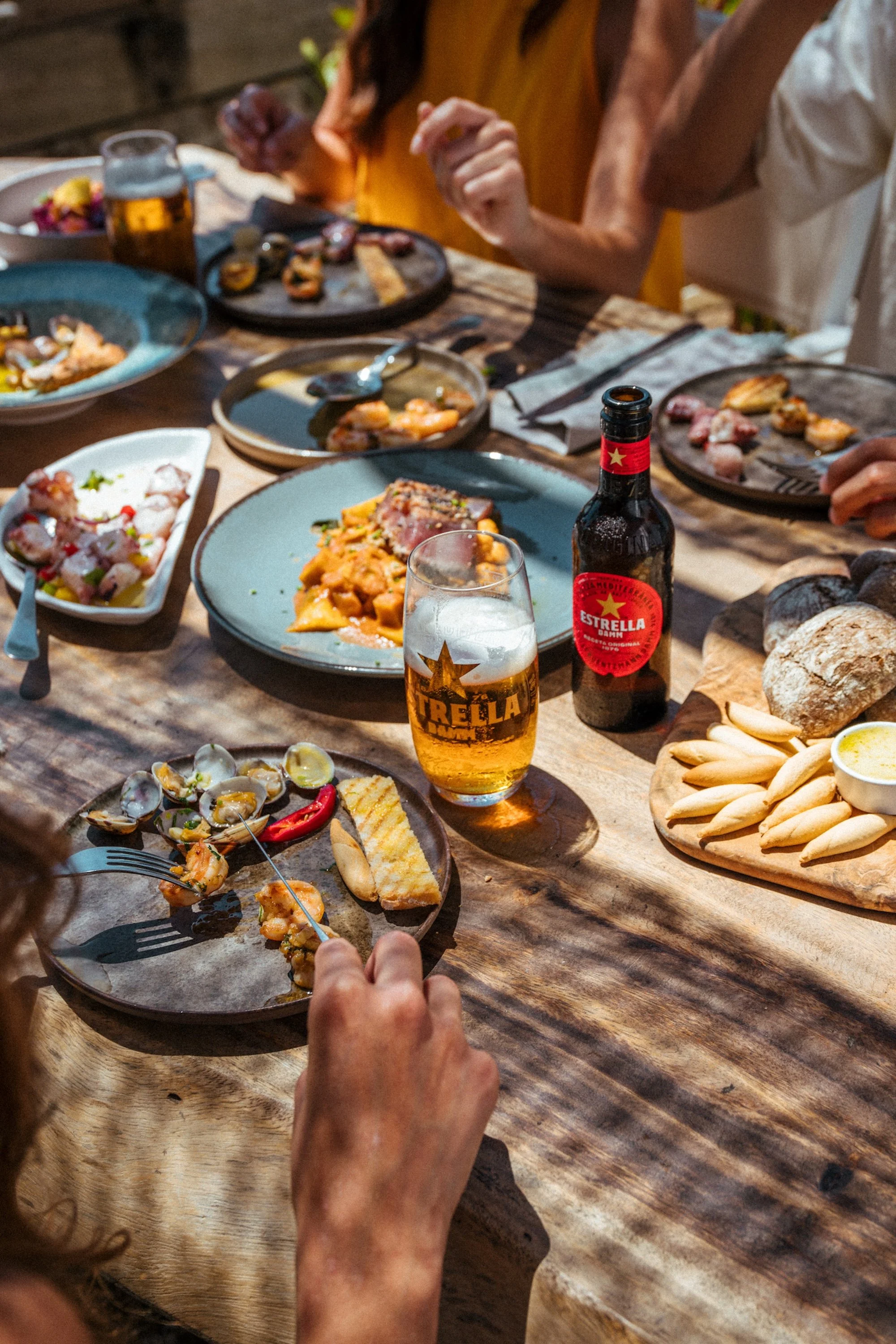 People enjoying a meal outdoors with various dishes, a glass of beer, and a bottle of Estrella beer on a rustic wooden table.