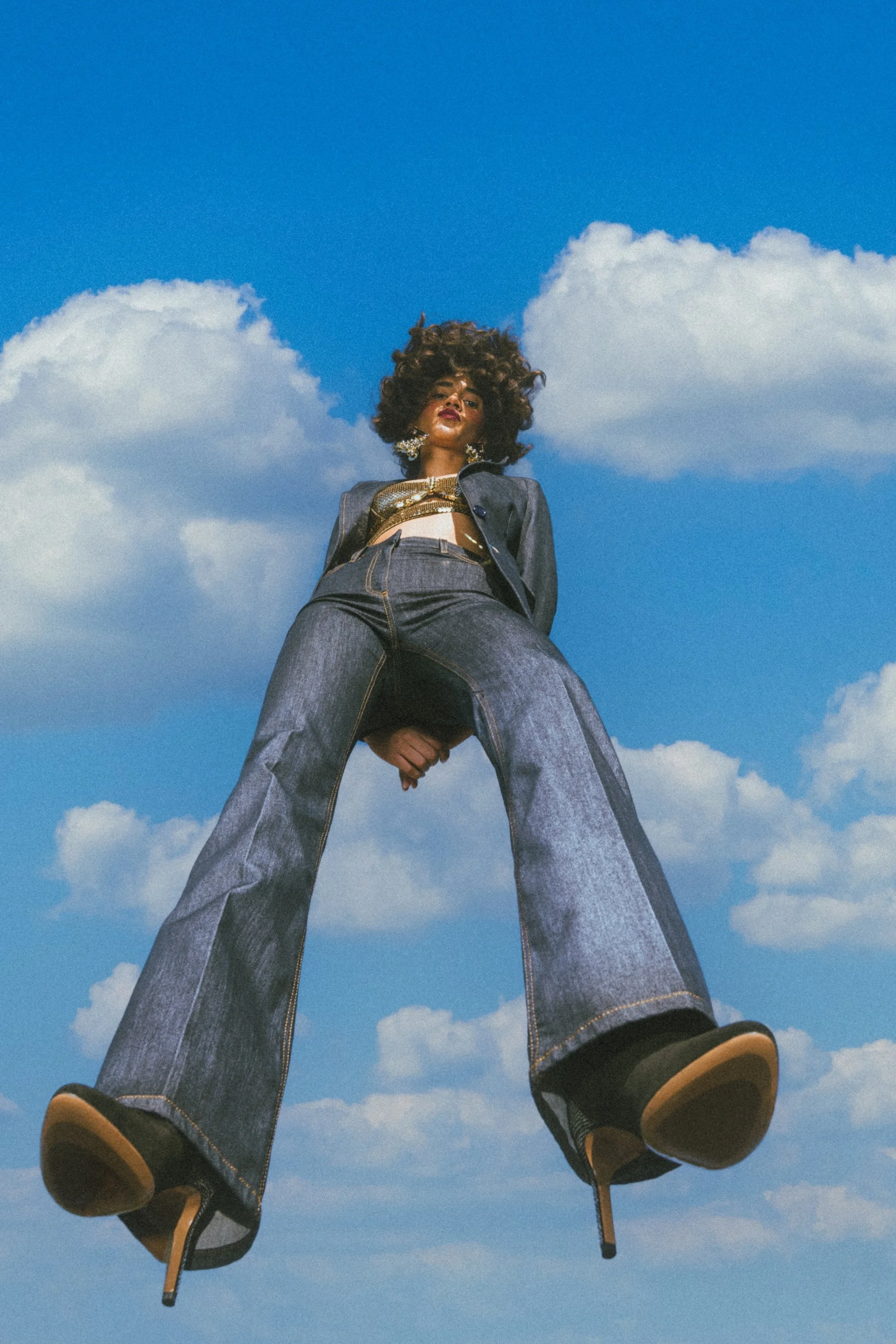 A woman with a large curly hairstyle standing against a bright blue sky with scattered white clouds, wearing jeans, a gold top, and a dark jacket, looking down at the camera with a confident expression.