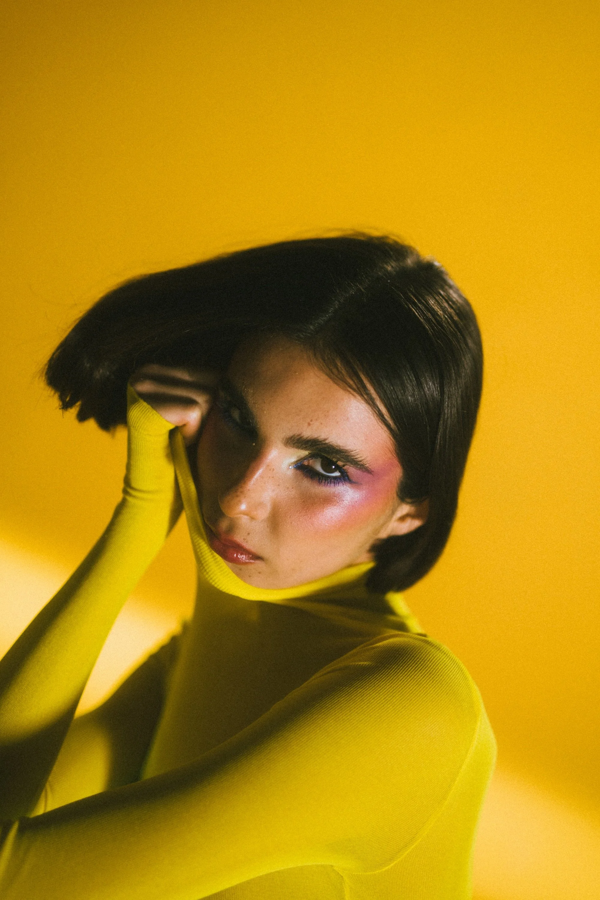 A woman with short black hair, wearing a yellow long-sleeve top, is posing with her hand near her face against a yellow background.