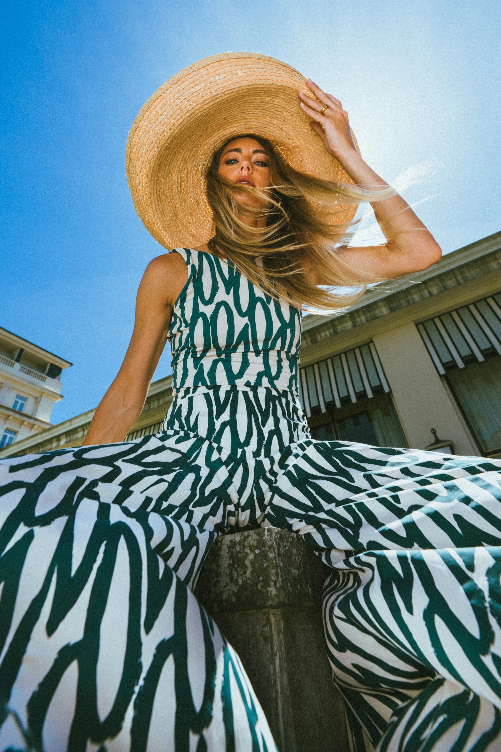 A woman wearing a wide-brimmed straw hat and a zebra print dress, sitting outdoors with a building and clear blue sky in the background.