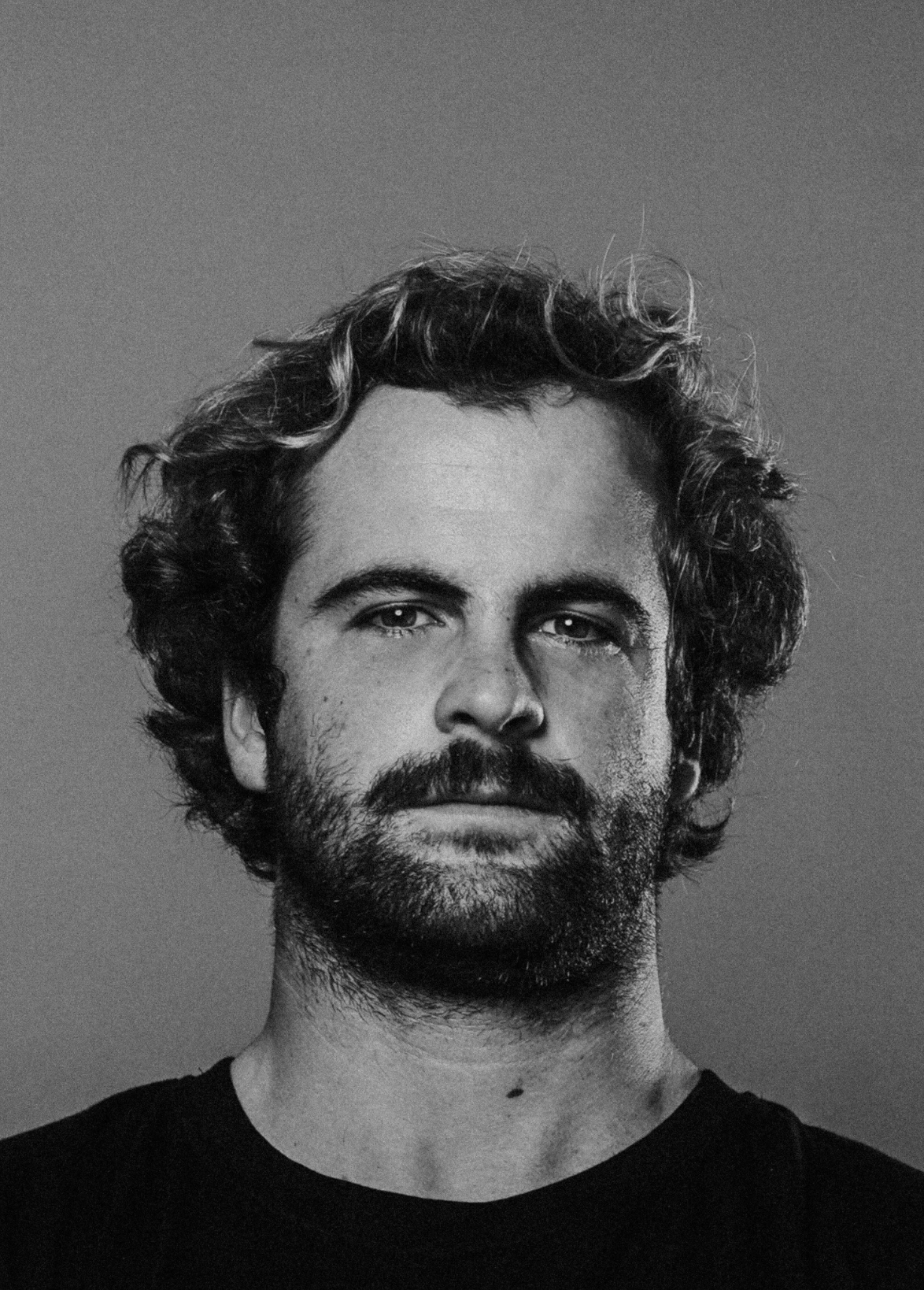 Black and white portrait of a man with curly hair and a beard, wearing a dark shirt, looking at the camera against a plain background.