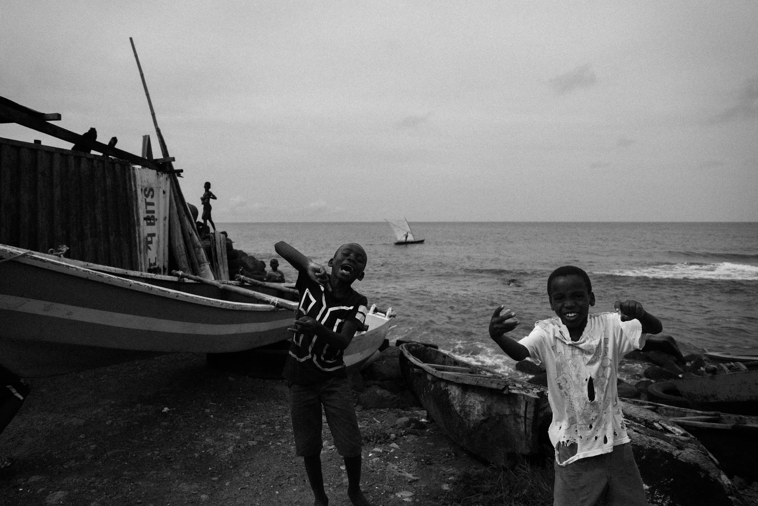 Black and white photo of children on a beach in front of boats, with a sailboat in the background on the water. Two children are making gestures towards the camera, smiling.