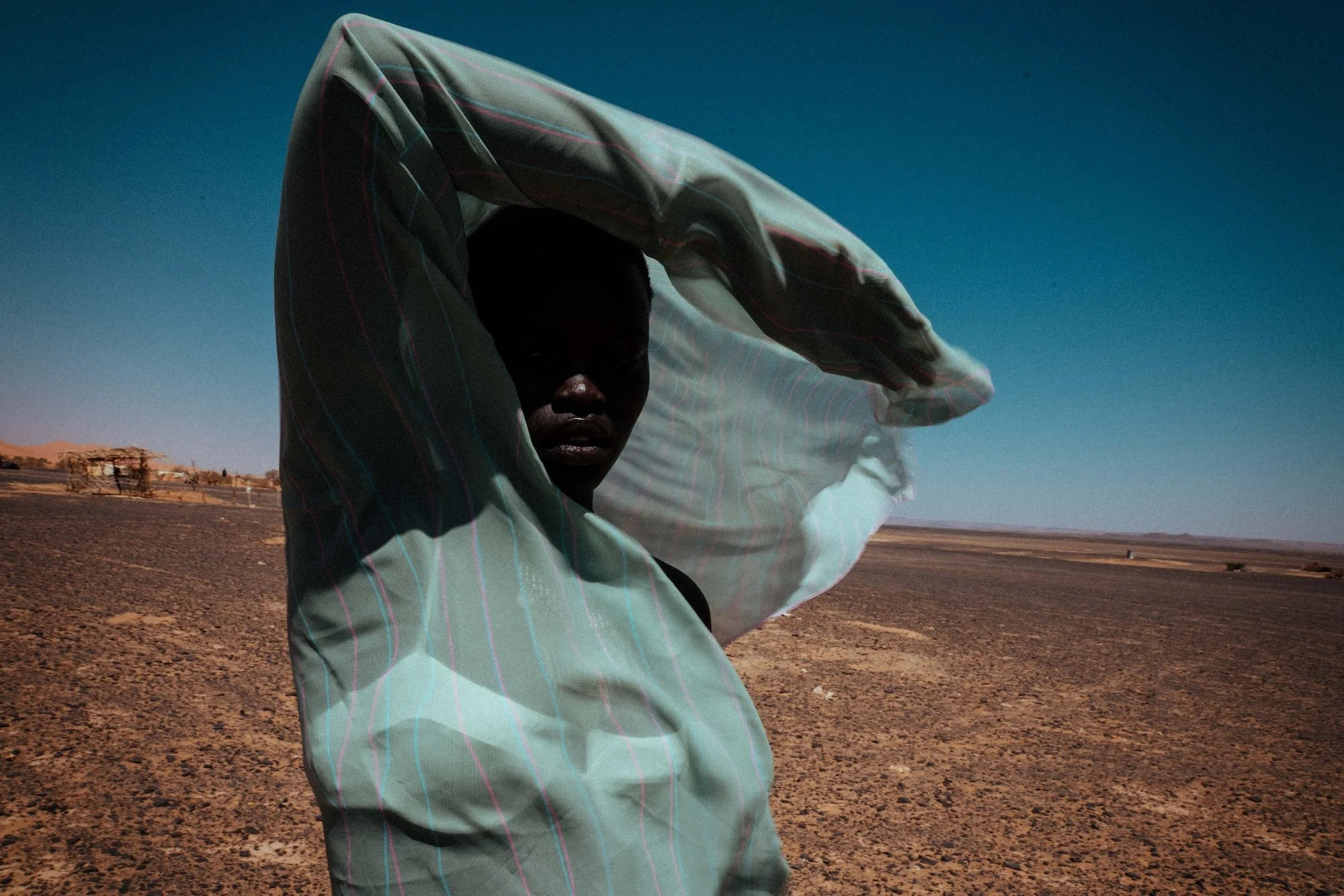 A person standing outdoors in a desert landscape with a clear blue sky, wearing a light-colored shirt and shielding their eyes from the sun with one hand.