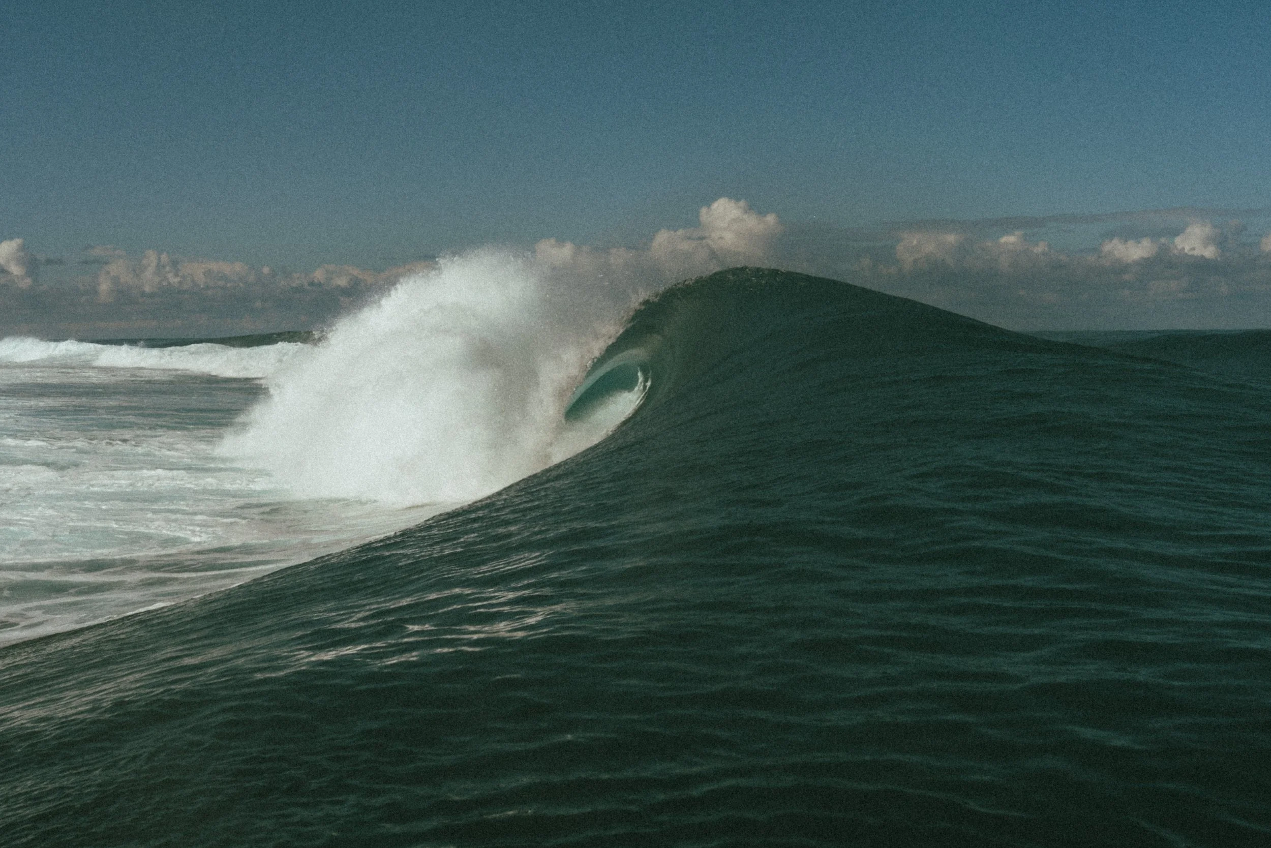 Ocean waves with a large wave curling over in the foreground and a distant mountain behind the wave, under a partly cloudy sky.
