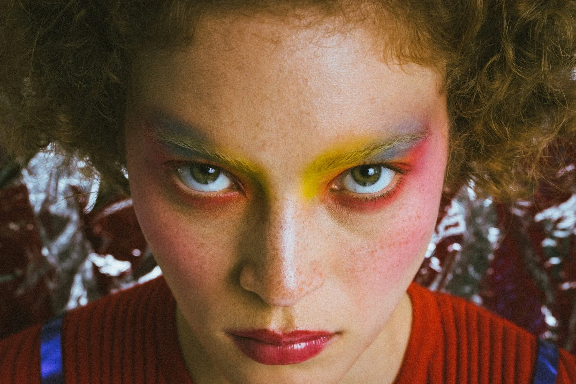 Close-up of a person's face with curly hair, colorful eye makeup, and a serious expression.