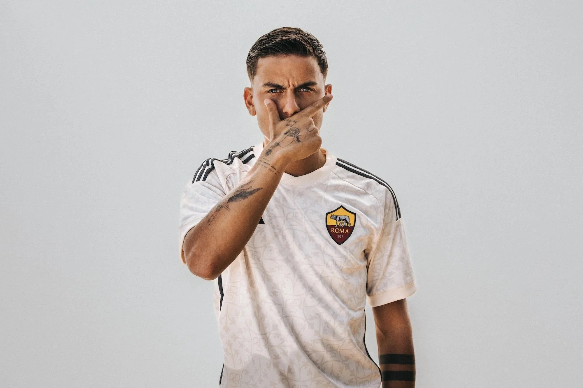 A young man with tattoos on his hand and arm, wearing a white soccer jersey with black stripes and the AS Roma logo, making a thoughtful gesture with his hand over his face against a plain light-colored background.