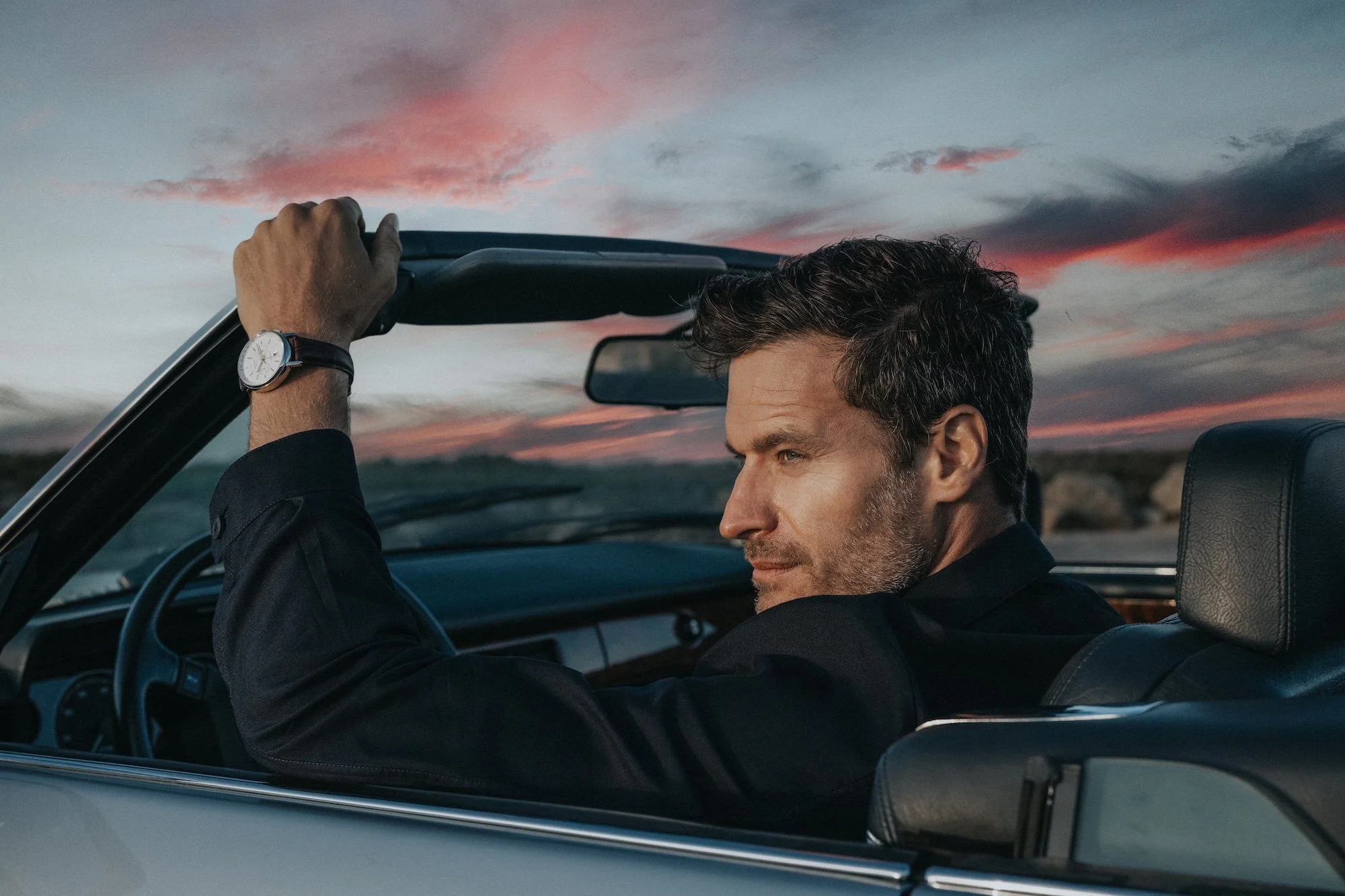 A man with dark hair and a beard driving a classic convertible car at sunset, with pink and gray clouds in the sky. He is wearing a black shirt and a watch.