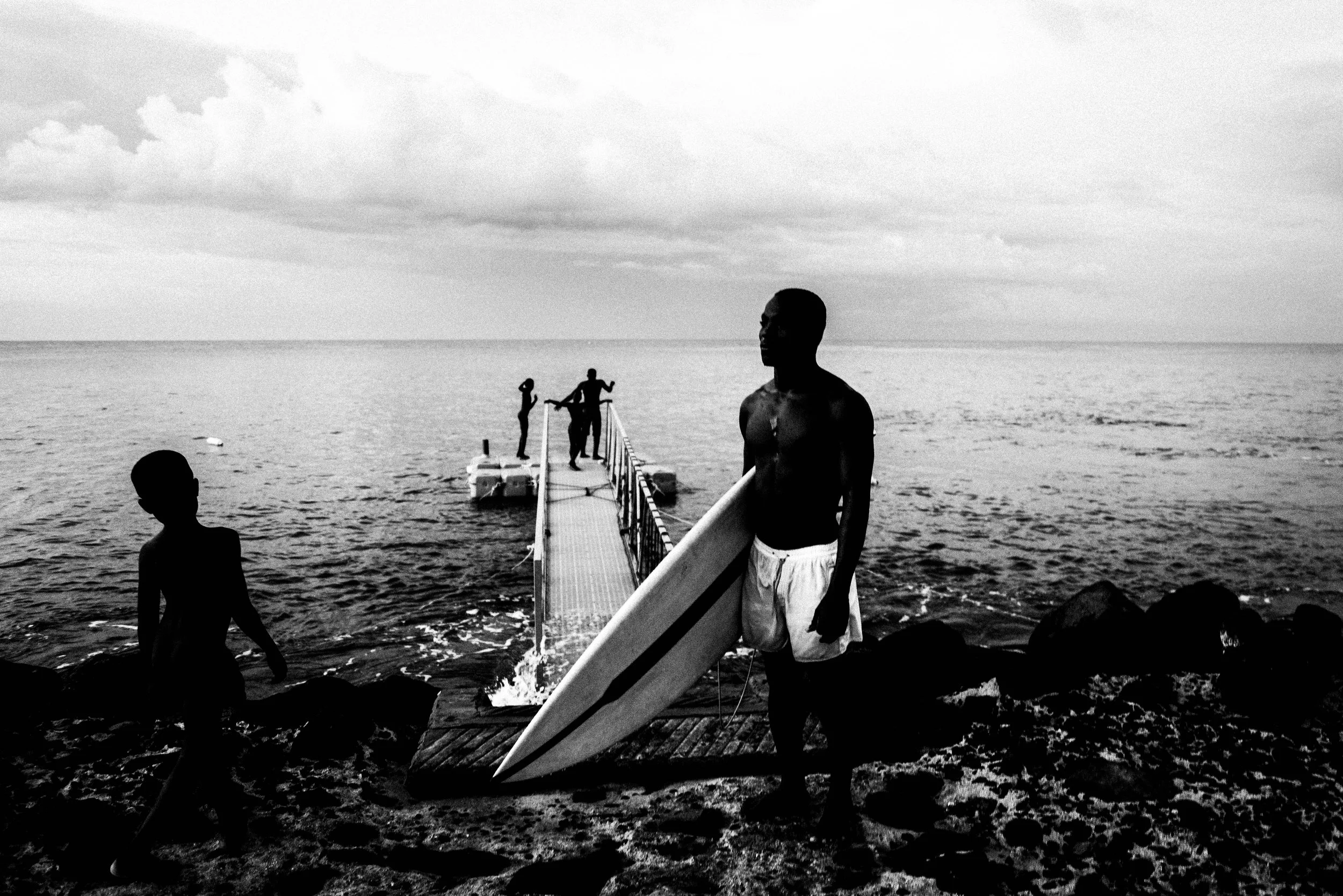 People at a beach with a dock extending into the water, some holding surfboards, under a cloudy sky.