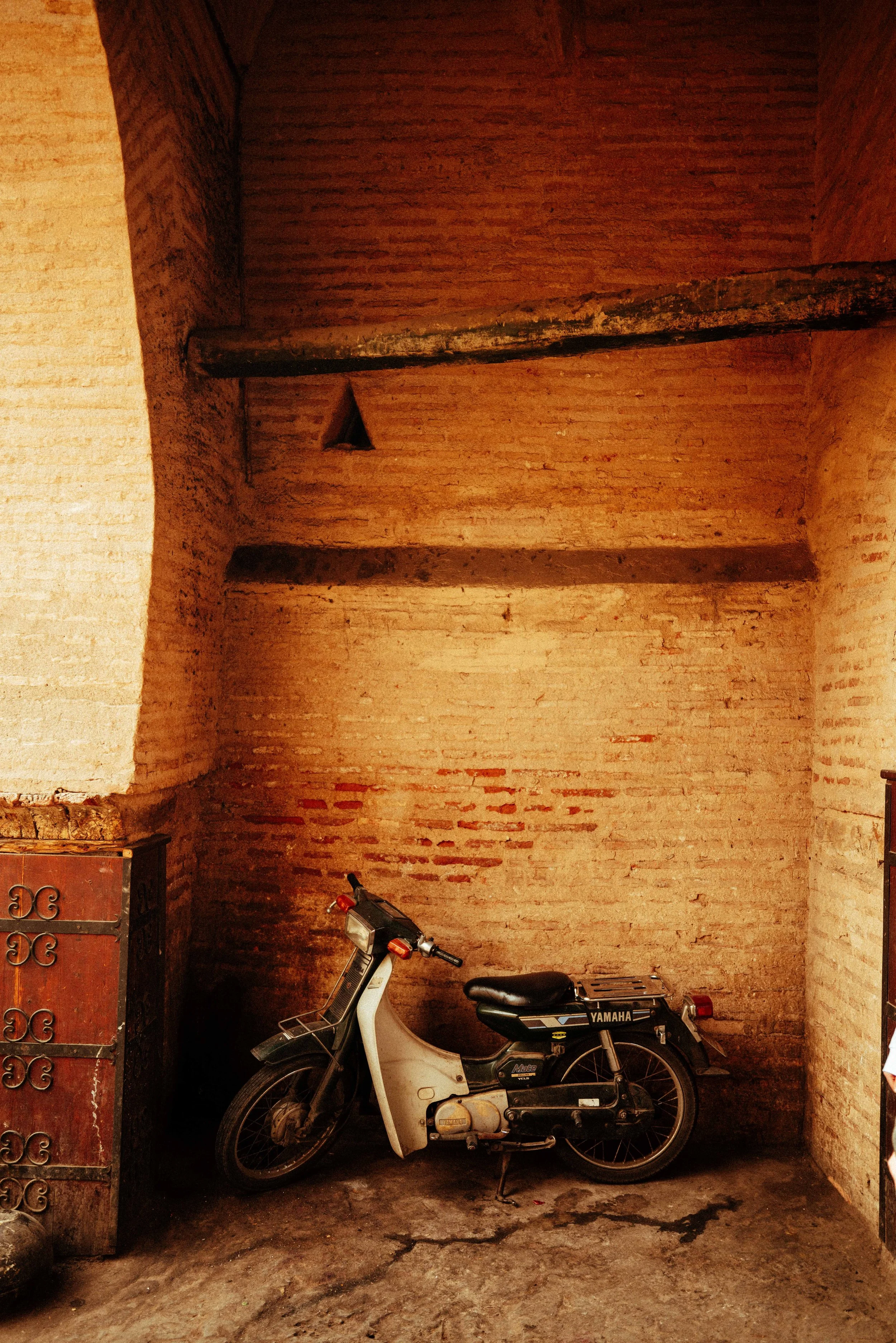 A vintage Yamaha motorcycle parked against a brick wall with a rough concrete floor, a small cabinet on the left, and a bicycle handlebar visible at the bottom right.