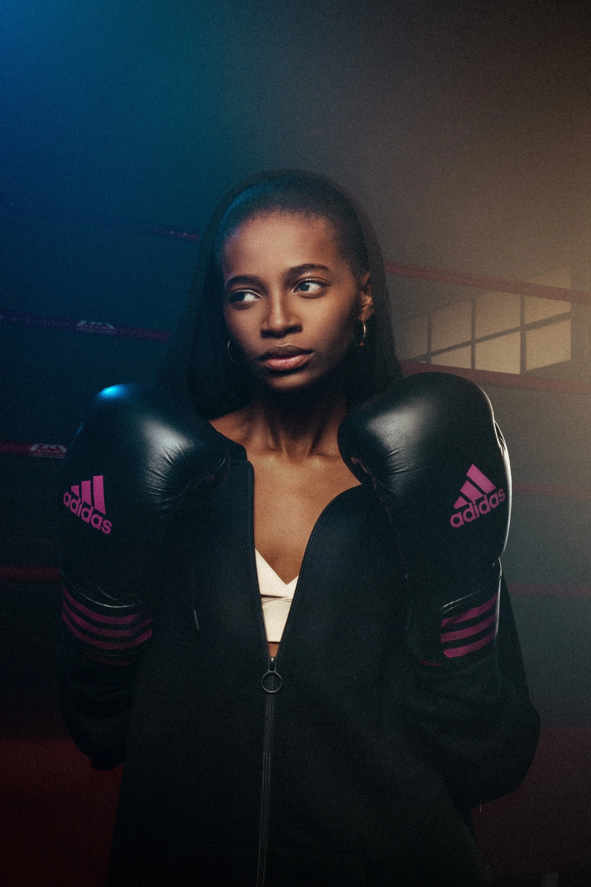 A woman with dark skin and straight black hair wearing boxing gloves and a black Adidas jacket with pink stripes, standing in a boxing gym with a boxing ring and wooden floor in the background.