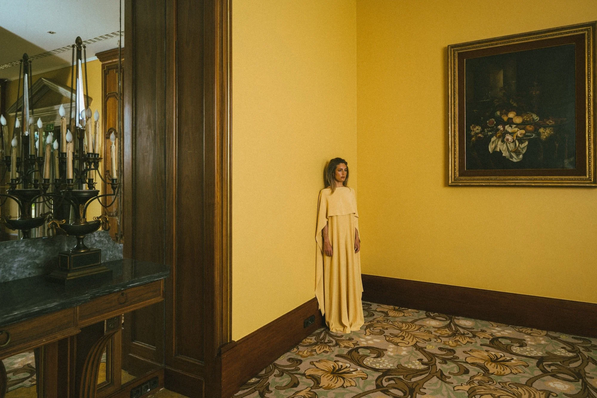 A woman in a yellow dress standing against a yellow wall in an elegant room with artwork and ornate decor.