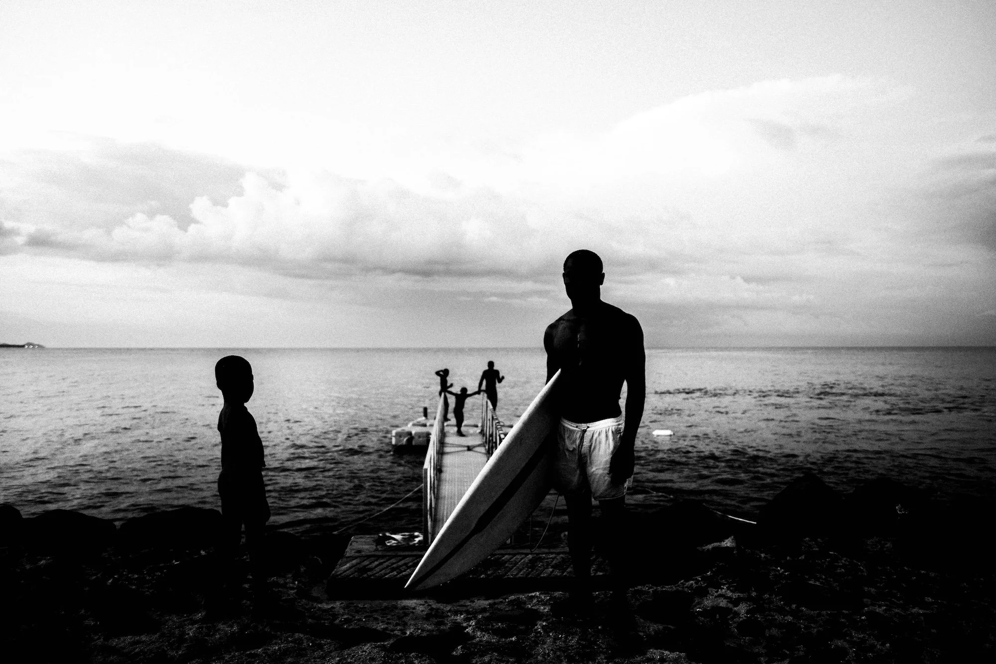 Silhouettes of people at the beach, including a person holding a surfboard, with a pier extending into the water and children playing, in black and white.
