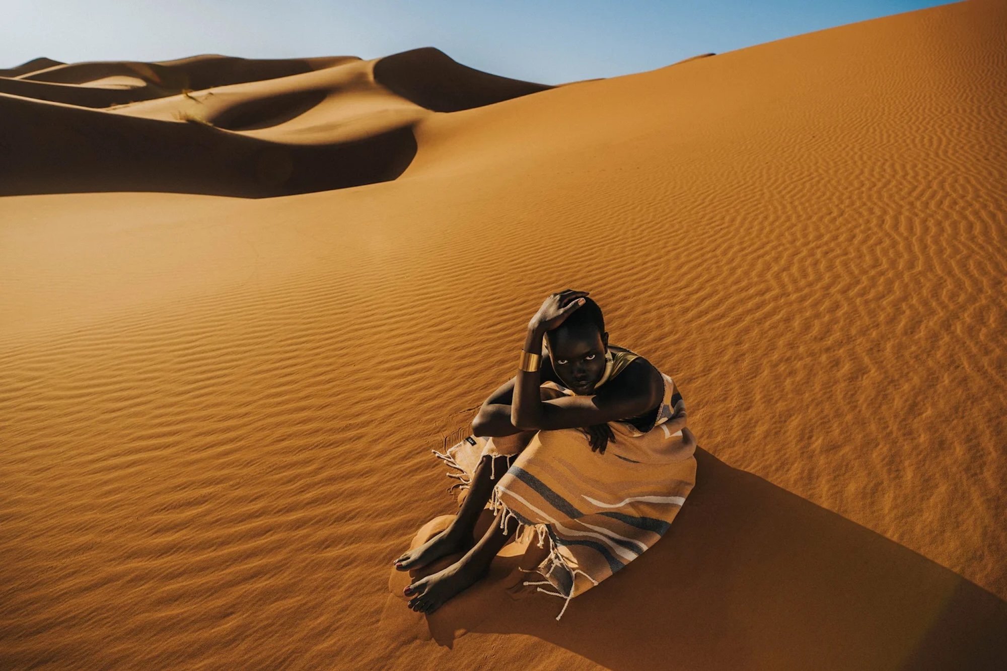 A woman with dark skin and short hair sitting on sand dunes in a desert. She is wearing a striped scarf, beige pants, and a long-sleeved shirt. She is leaning forward with one arm resting on her knee, and her other hand on her head, looking directly 