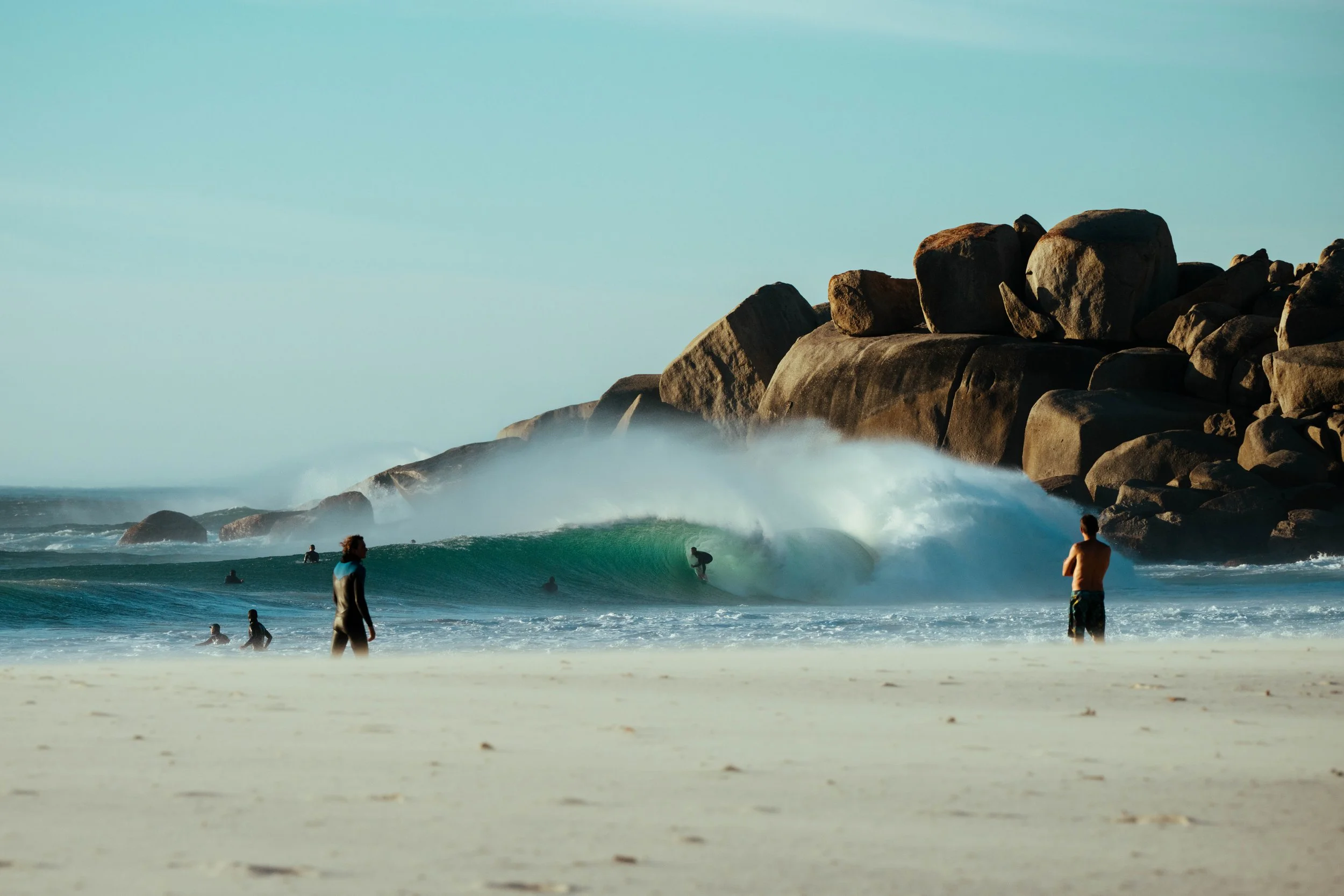 People surfing and swimming in the ocean near rocky cliffs with waves crashing onto the shore.