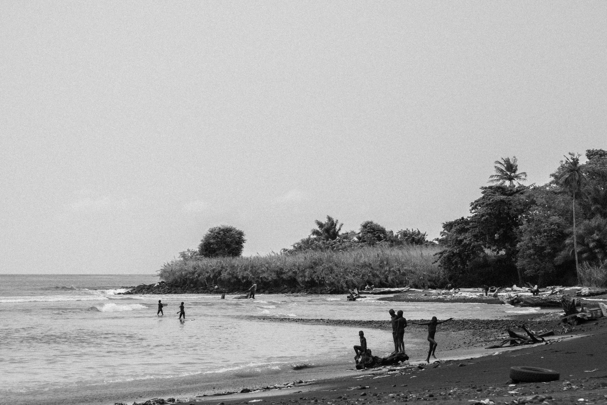 Black and white photo of a beach scene with people playing near the shoreline, trees and shrubs on a small island or peninsula in the background.