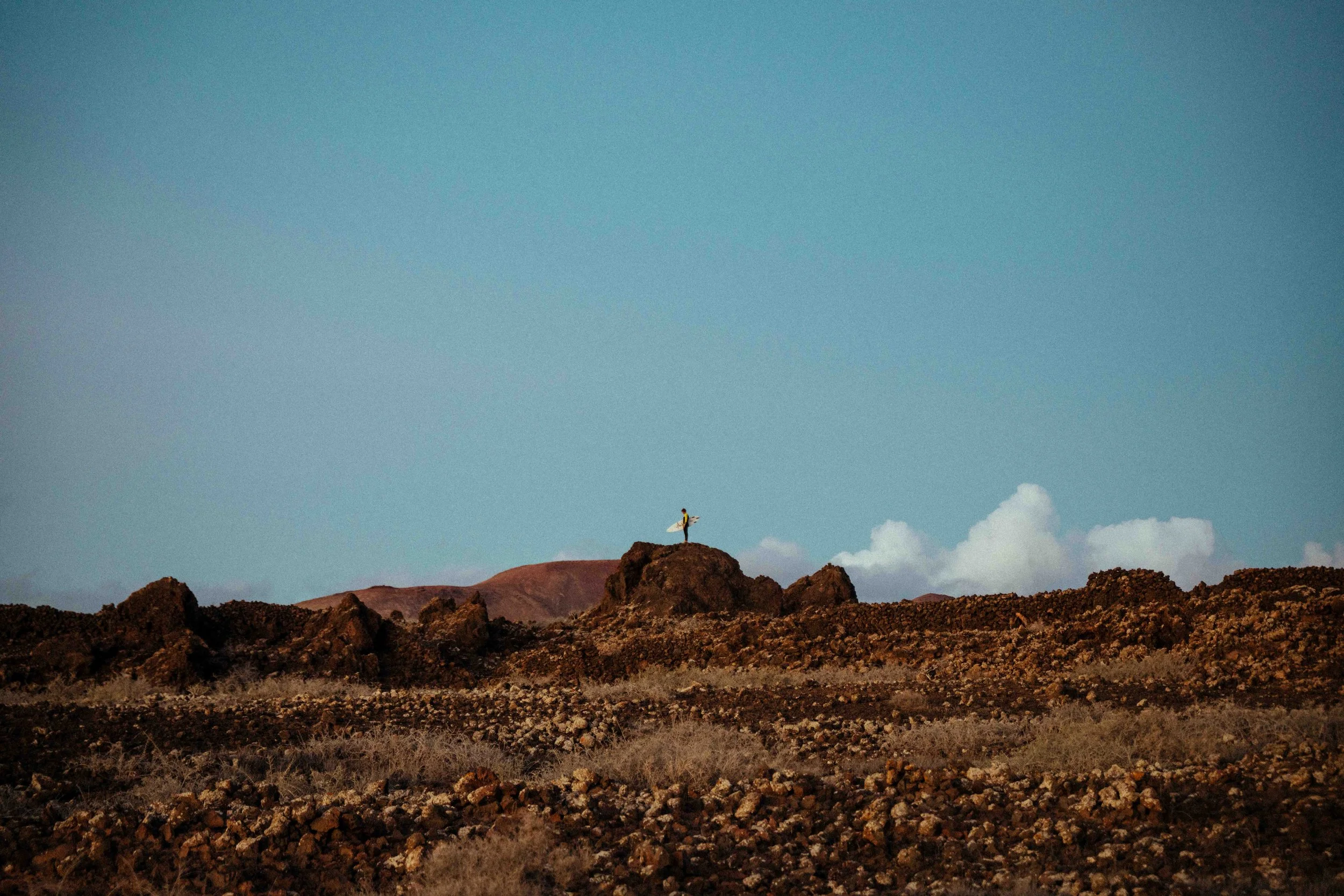 A person standing on a rocky hill with arms raised, holding a surfboard, in a desert landscape with mountains and a blue sky with clouds.