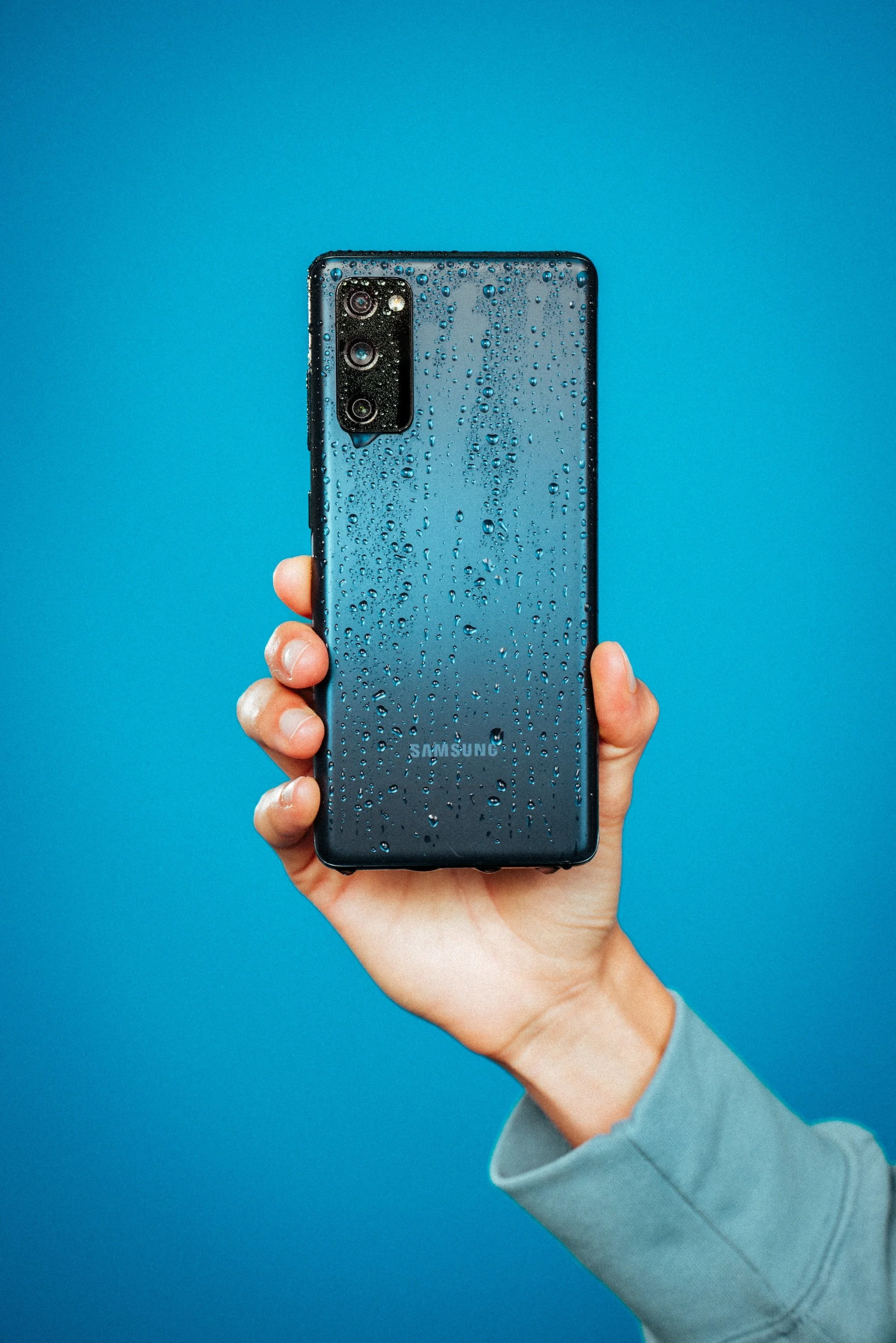 A person holding a Samsung smartphone with water droplets on the back, against a solid blue background.