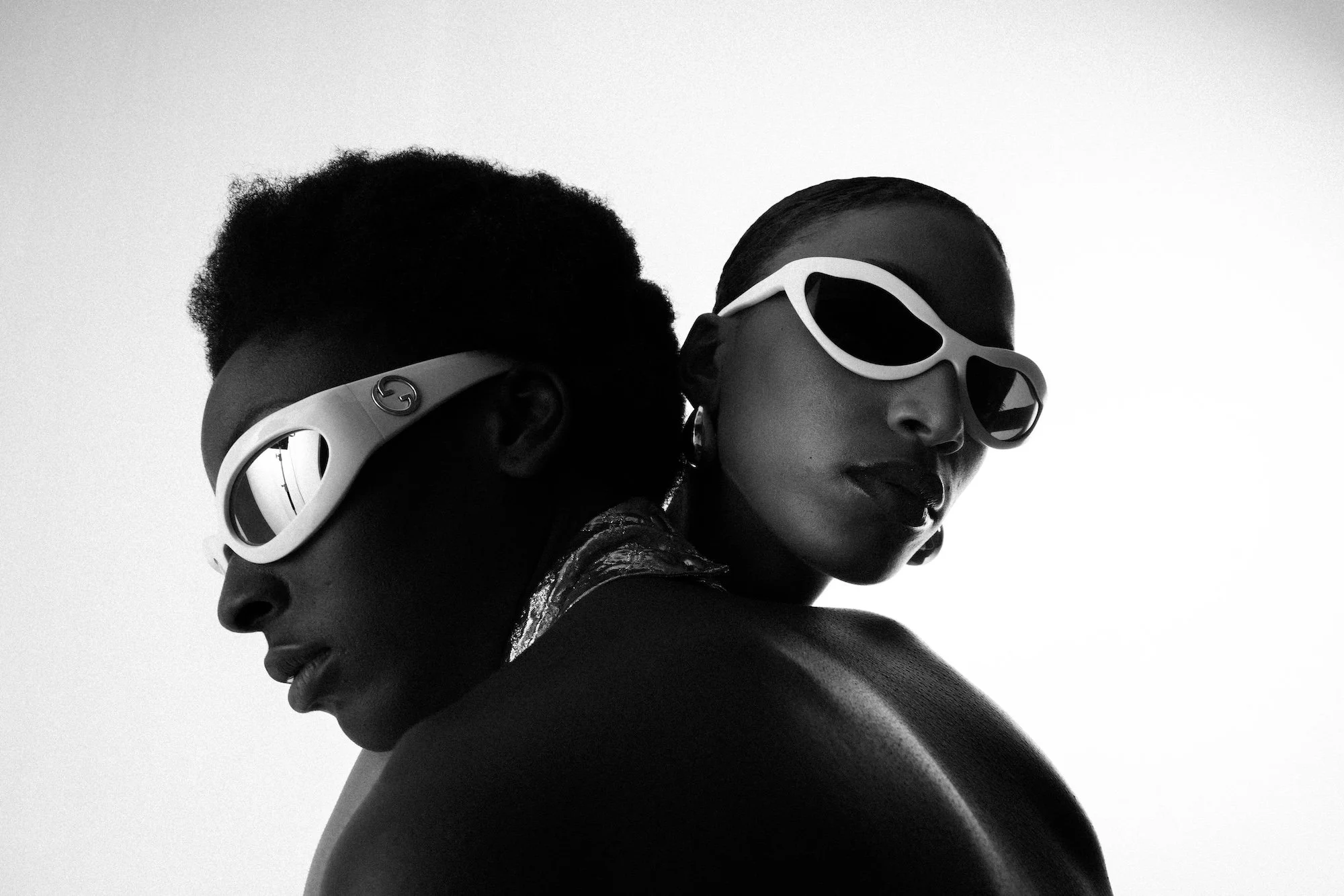 Black and white photo of two women with contrasting skin tones wearing futuristic sunglasses, facing opposite directions and closely positioned, with their faces and shoulders partially visible against a plain background.